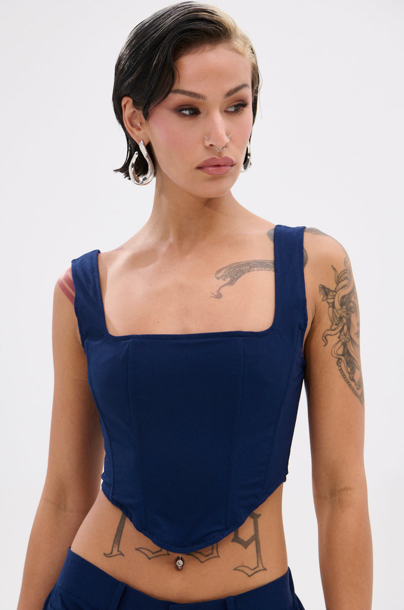 A person with short, dark, side-parted hair models the FEELING GOOD CORSET in navy blue with a square neckline. Arm and chest tattoos and facial piercings complete their look as they gaze slightly to the side.