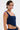 A person with short dark hair, hoop earrings, and visible upper arm and torso tattoos poses against a plain background, wearing the FEELING GOOD CORSET—a sleeveless navy blue top with a square neckline.