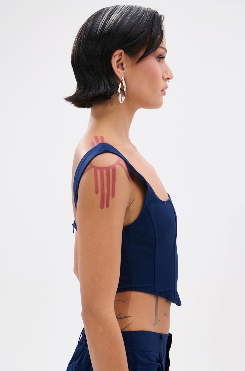 A person with short dark hair and bold pink makeup stands in profile, wearing the FEELING GOOD CORSET with a square neckline. They have large hoop earrings, red lines on their shoulder, and visible tattoos on their arm and side.