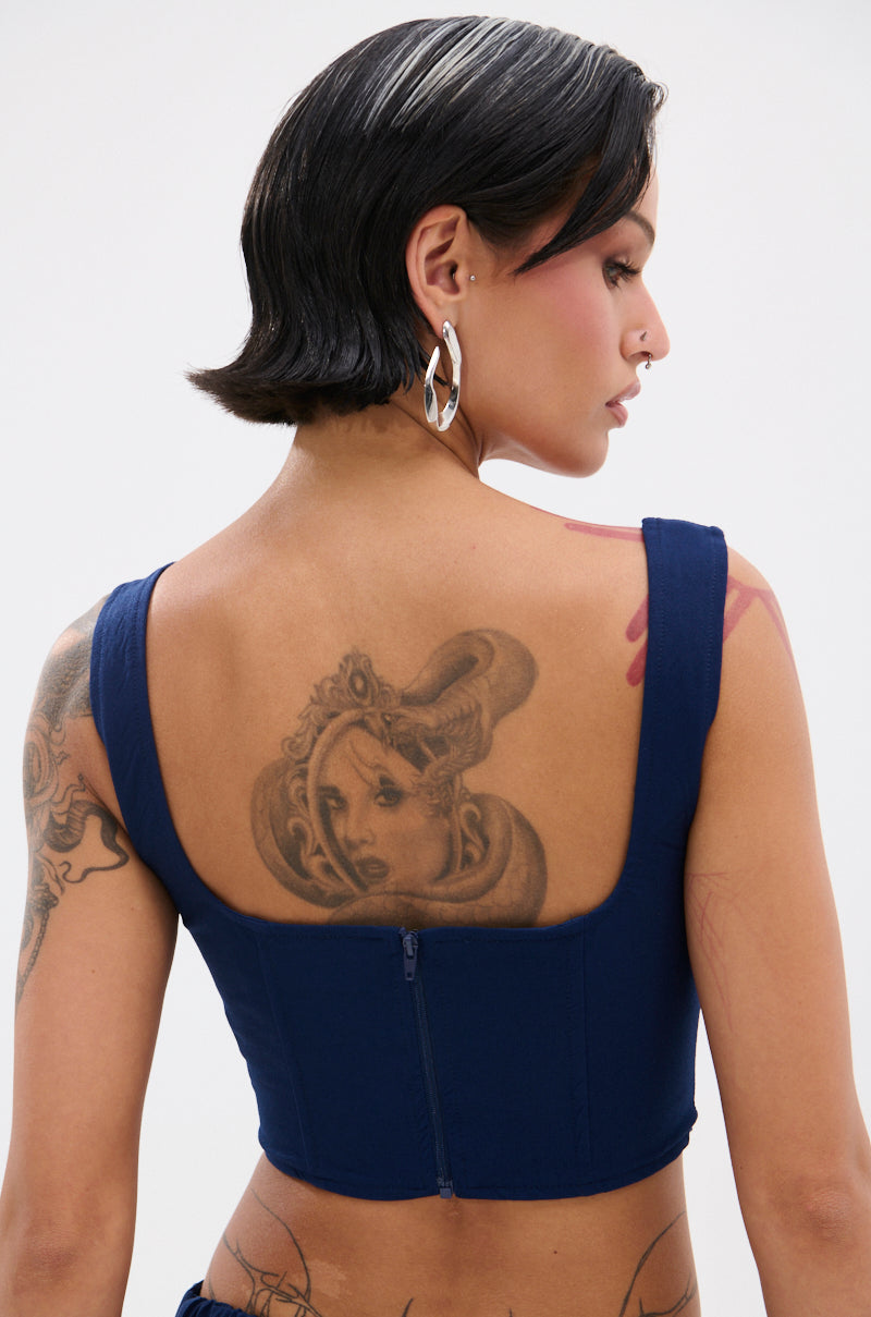 A woman with short dark hair and silver earrings is shown from the back, wearing the FEELING GOOD CORSET—a sleeveless blue top with a square neckline in sleek, lightweight fabric—revealing bold upper back tattoo art.