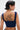 A woman with short dark hair and silver earrings is shown from the back, wearing the FEELING GOOD CORSET—a sleeveless blue top with a square neckline in sleek, lightweight fabric—revealing bold upper back tattoo art.