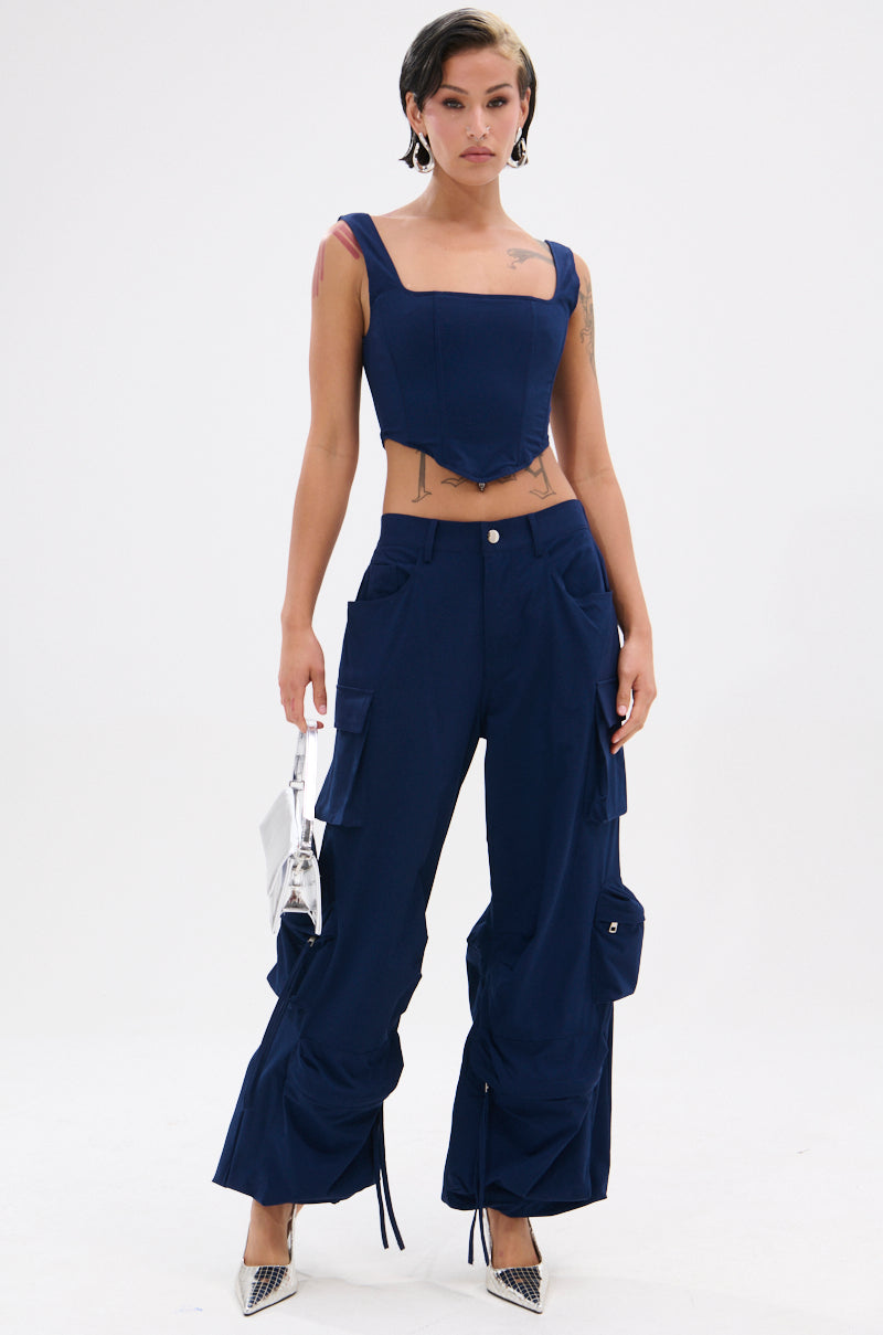 A woman stands against a white background wearing the FEELING GOOD CORSET, a navy blue fitted top with a square neckline, and matching cargo pants with pockets and drawstrings. She accessorizes with a silver handbag and pointed silver heels.