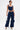 A woman stands against a white background wearing the FEELING GOOD CORSET, a navy blue fitted top with a square neckline, and matching cargo pants with pockets and drawstrings. She accessorizes with a silver handbag and pointed silver heels.