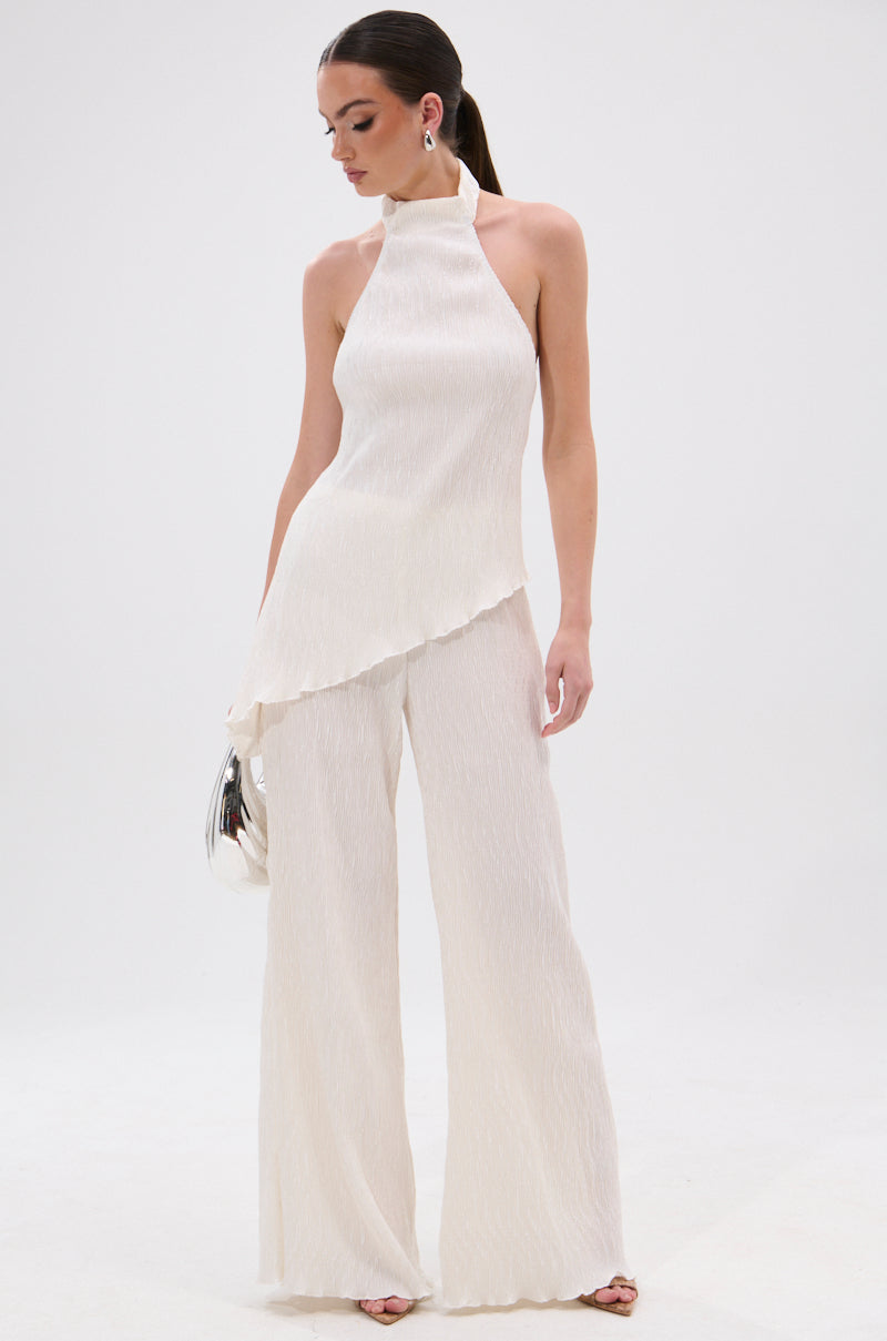 A woman stands against a plain white background wearing a sleeveless, high-neck, off-white asymmetrical top and OLIVIA PLISSE PANT IN WHITE featuring a wide-leg silhouette. She holds a small silver clutch with her hair in a sleek ponytail. 0