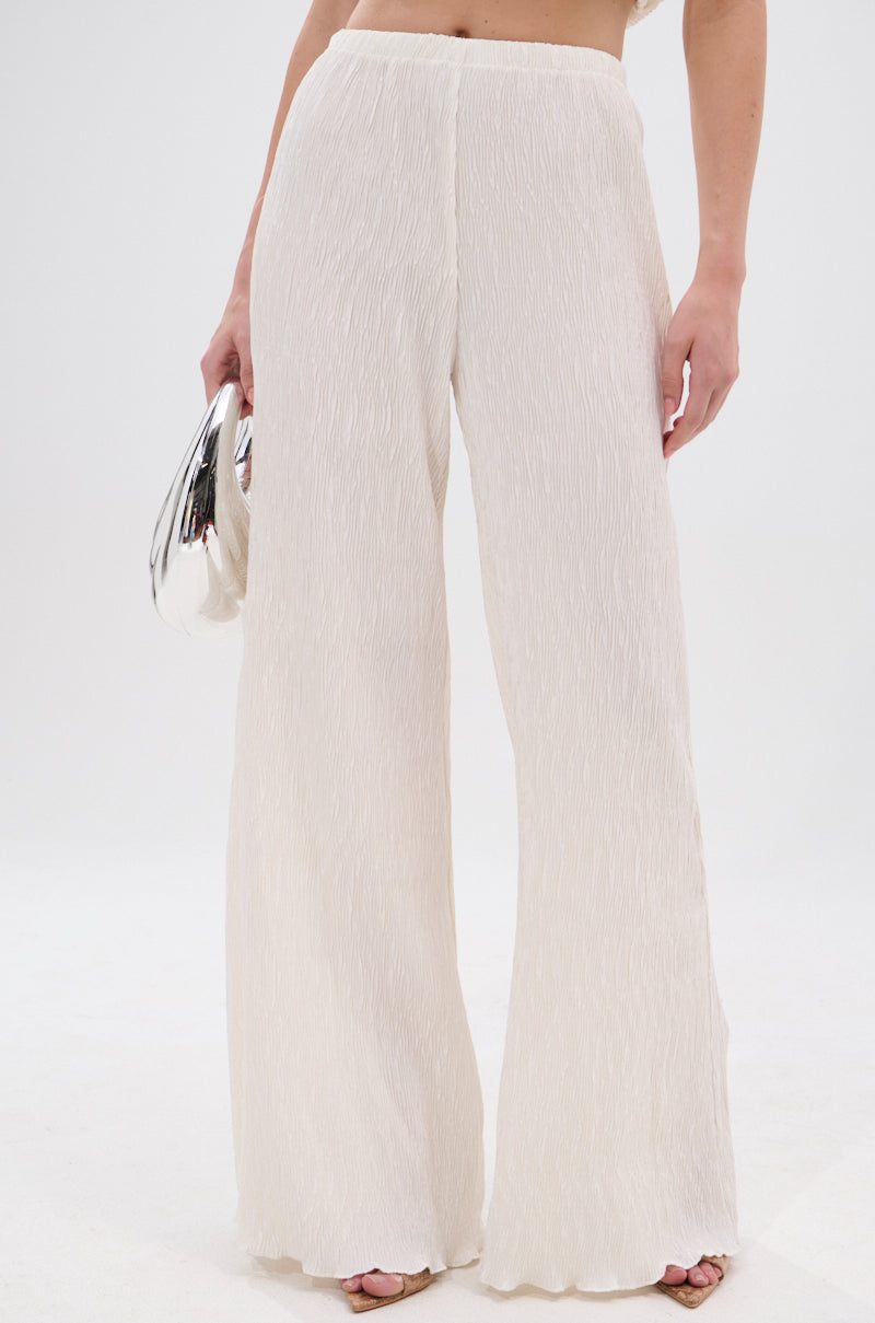 Wearing the OLIVIA PLISSE PANT IN WHITE with a high-rise fit and wide-leg silhouette, a person stands against a white background, holding a small metallic silver clutch in their left hand. 1