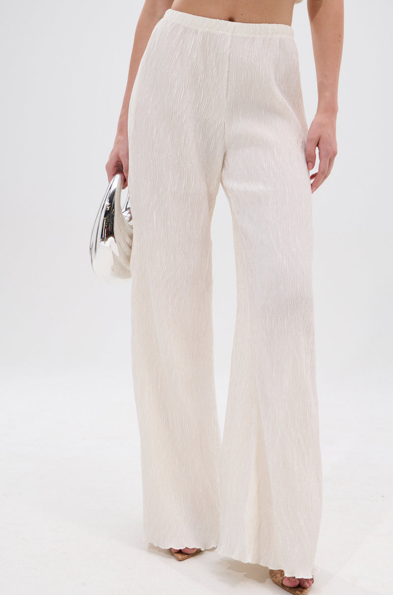 Someone wears the OLIVIA PLISSE PANT IN WHITE, featuring a high-rise fit and wide-leg silhouette, styled with a shiny silver clutch against a plain white background. 2