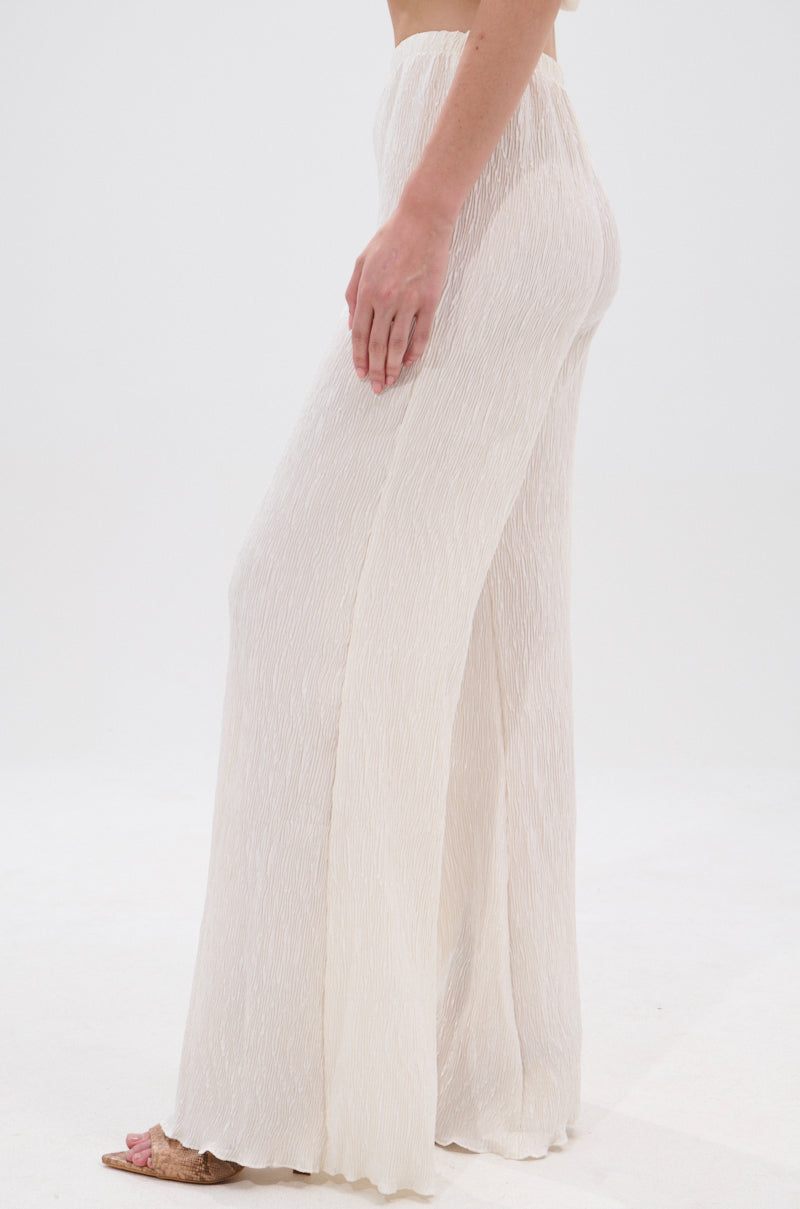 A person stands barefoot, modeling the OLIVIA PLISSE PANT IN WHITE. These high-rise, wide-leg textured pants are styled from the side against a plain white background. 3