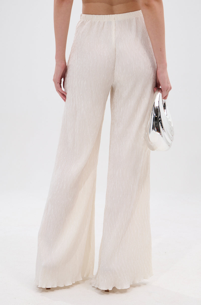 Wearing the OLIVIA PLISSE PANT IN WHITE with a wide-leg silhouette, a person stands facing away, holding a shiny silver clutch. The light-colored pants and background create a clean, minimalist vibe. 4