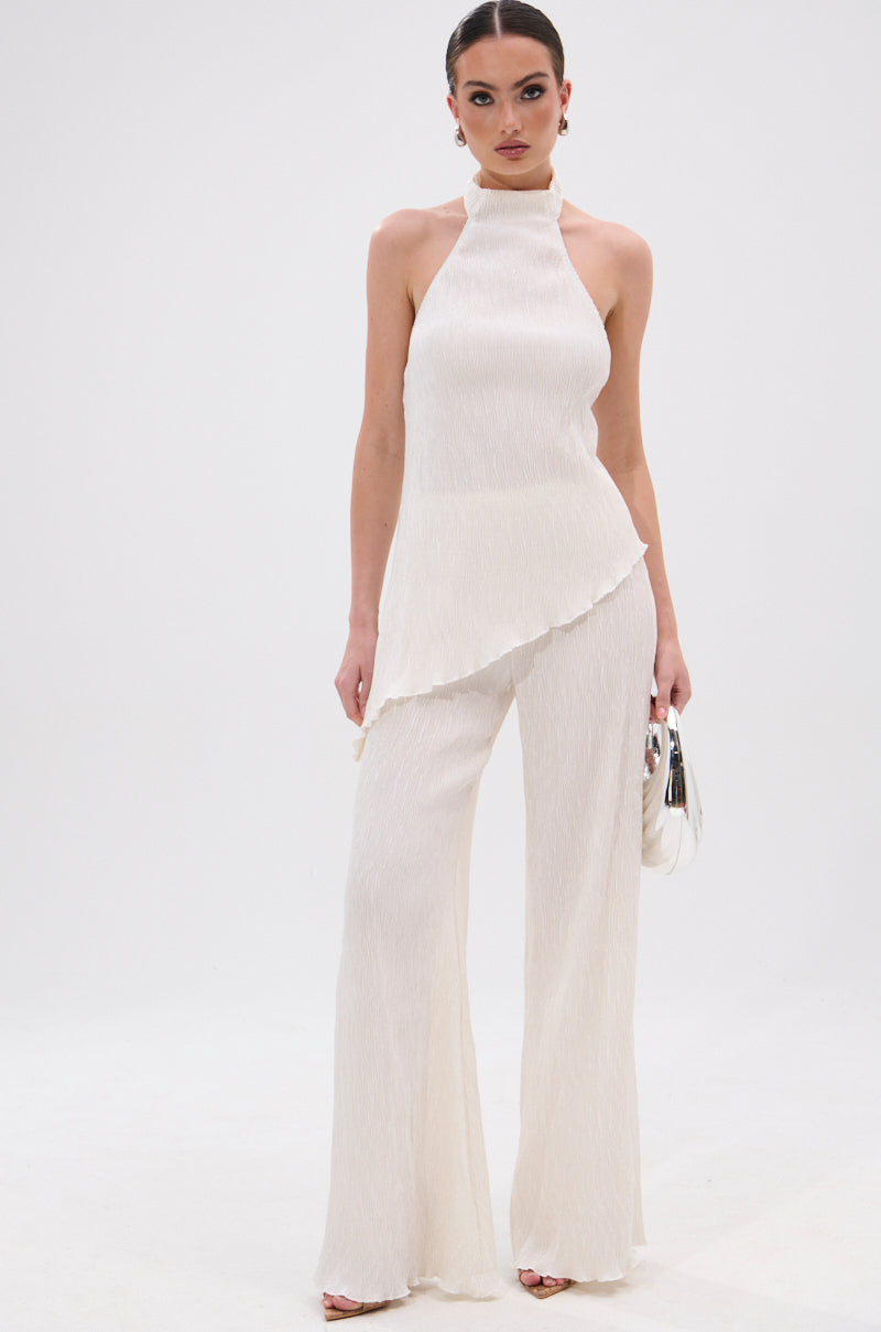 A woman stands against a plain white background, wearing the OLIVIA PLISSE PANT IN WHITE styled with a high-rise fit and wide-leg silhouette. She holds a small white handbag and has her hair in a sleek bun. 6
