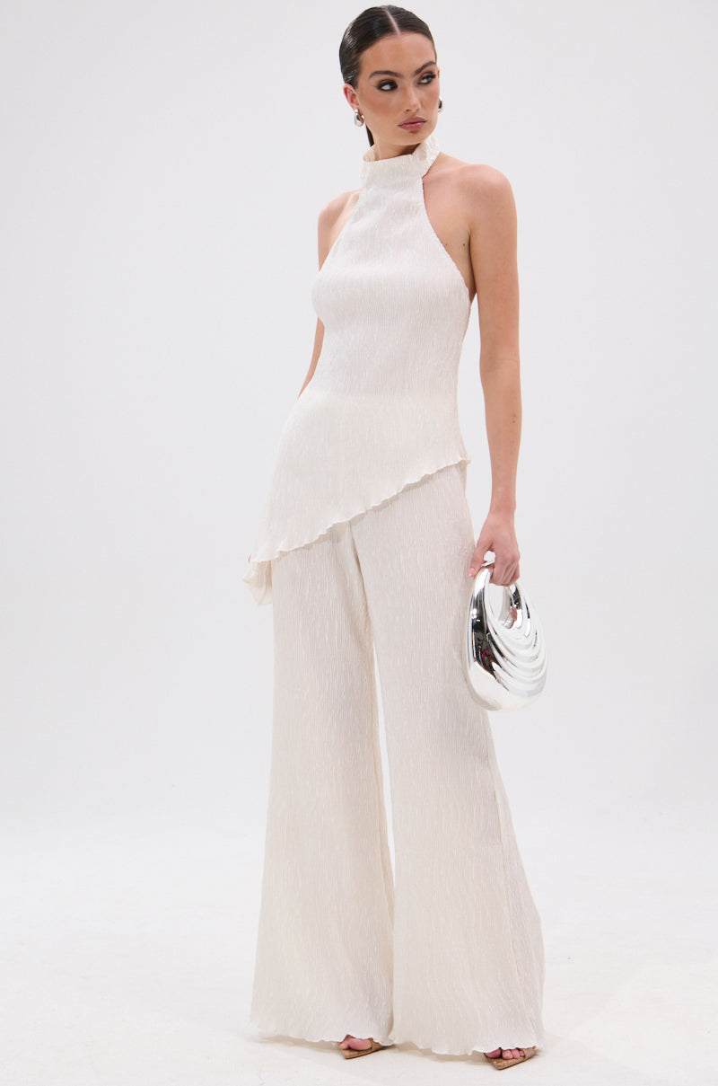 A woman poses against a white background wearing the OLIVIA PLISSE PANT IN WHITE, an elegant wide-leg, high-waisted style, paired with a sleeveless cream jumpsuit and a shiny silver handbag. 7