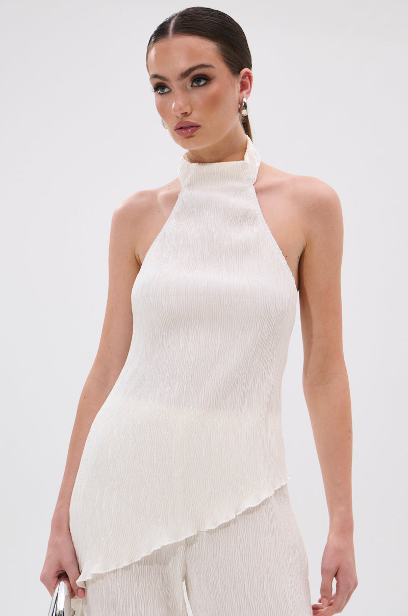 A woman stands against a plain white background, wearing the OLIVIA PLISSE HALTER NECK ASYMMETRICAL TOP IN WHITE, paired with matching white pants.