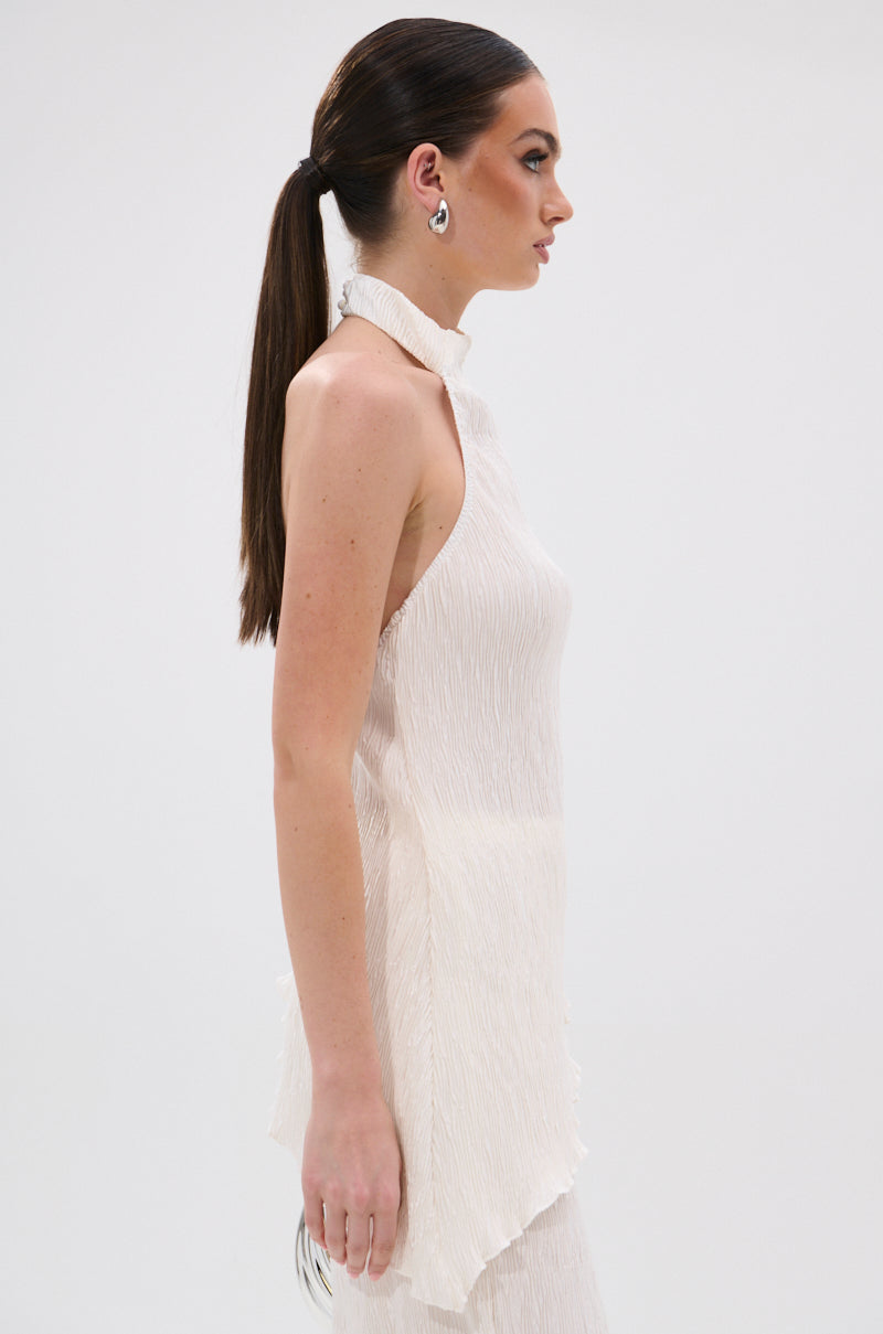 A woman with long brown hair in a low ponytail wears the OLIVIA PLISSE HALTER NECK ASYMMETRICAL TOP IN WHITE. She stands in profile against a plain white background, accessorized with silver hoop earrings.