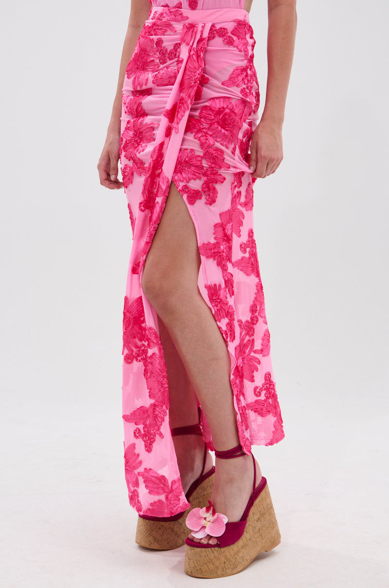 A person wearing the FLOWER GIRL MESH MAXI SKIRT, a pink floral maxi skirt with a high slit, paired with matching pink platform wedge sandals featuring flower details. The background is plain white.
