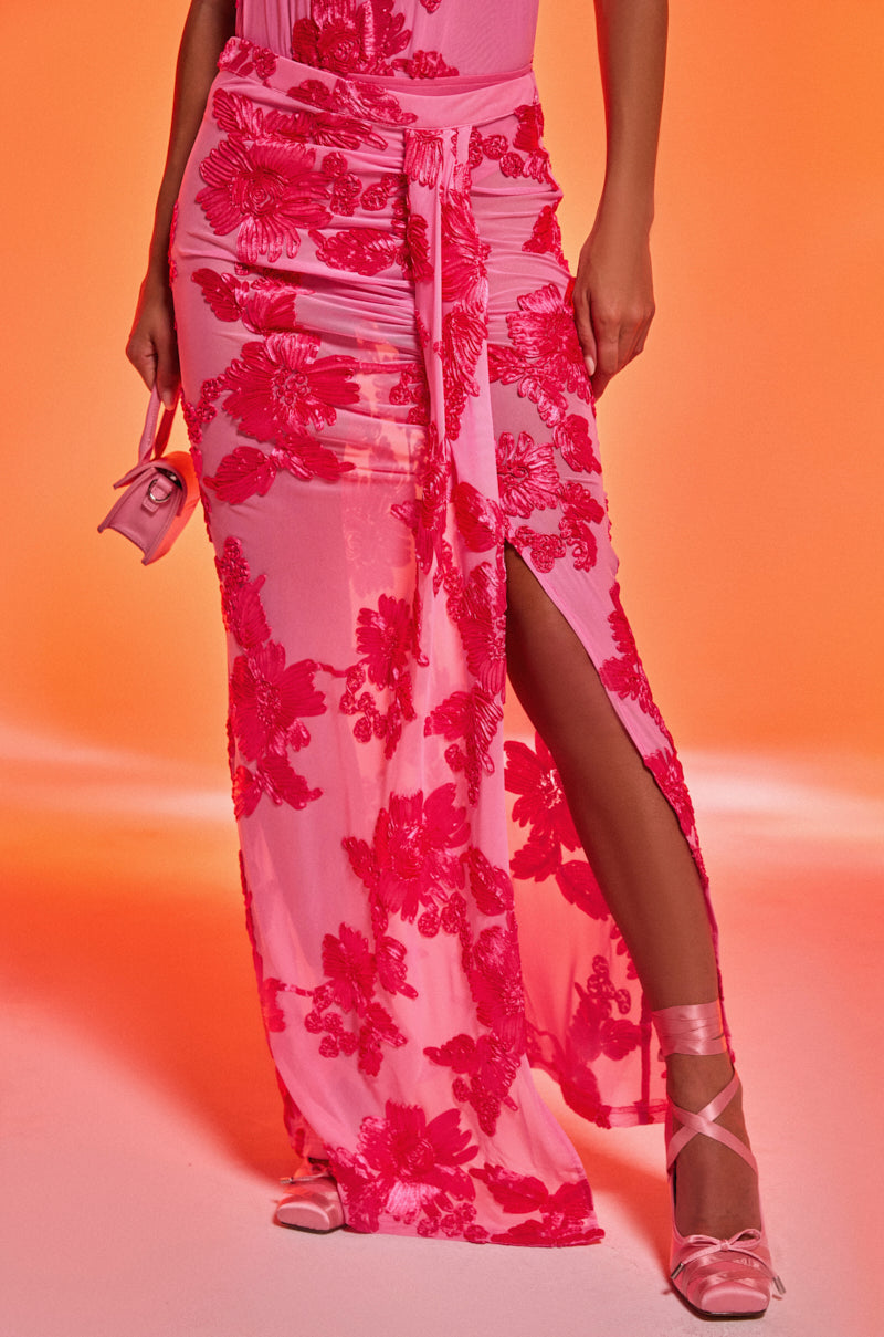 Wearing the FLOWER GIRL MESH MAXI SKIRT, featuring bold red florals and a thigh-high slit, this person pairs it with pink ballet flats, matching socks, and a small pink purse against an orange background.