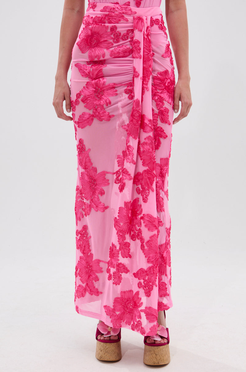 Wearing the FLOWER GIRL MESH MAXI SKIRT with bold red florals, wrap style, and tie waist, paired with red platform sandals. The sheer pink mesh adds elegance against a plain white background.