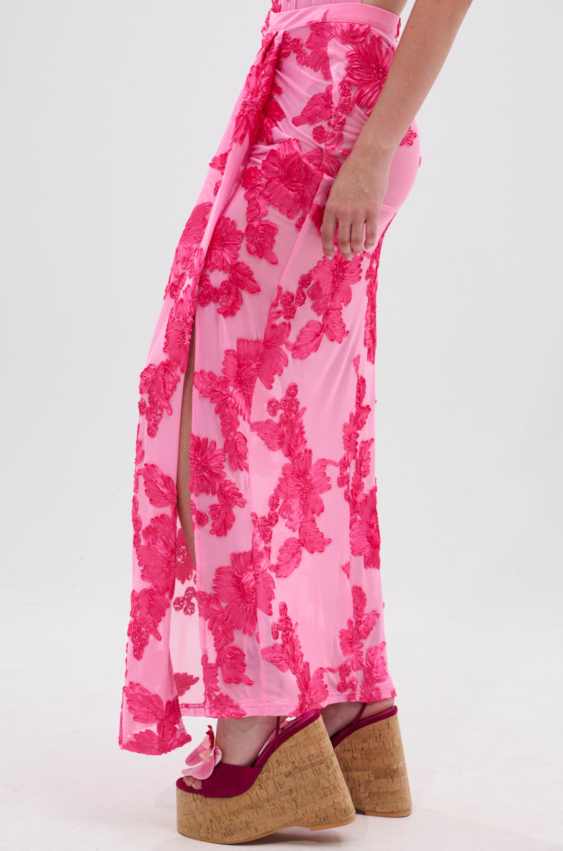 A person wears the FLOWER GIRL MESH MAXI SKIRT in pink with bold floral patterns and a high slit, paired with pink platform wedge sandals with cork soles. The background is plain white.