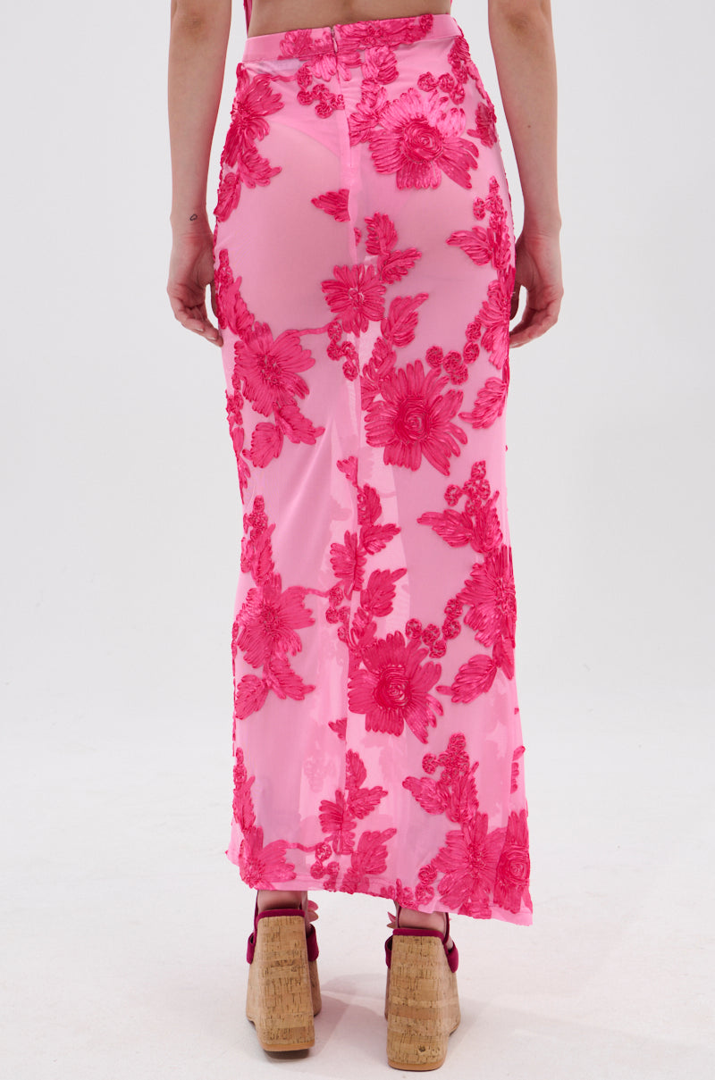 A person stands with their back to the camera, wearing the FLOWER GIRL MESH MAXI SKIRT featuring bright pink satin-textured floral appliqués and a high-rise fit, paired with tan platform sandals.