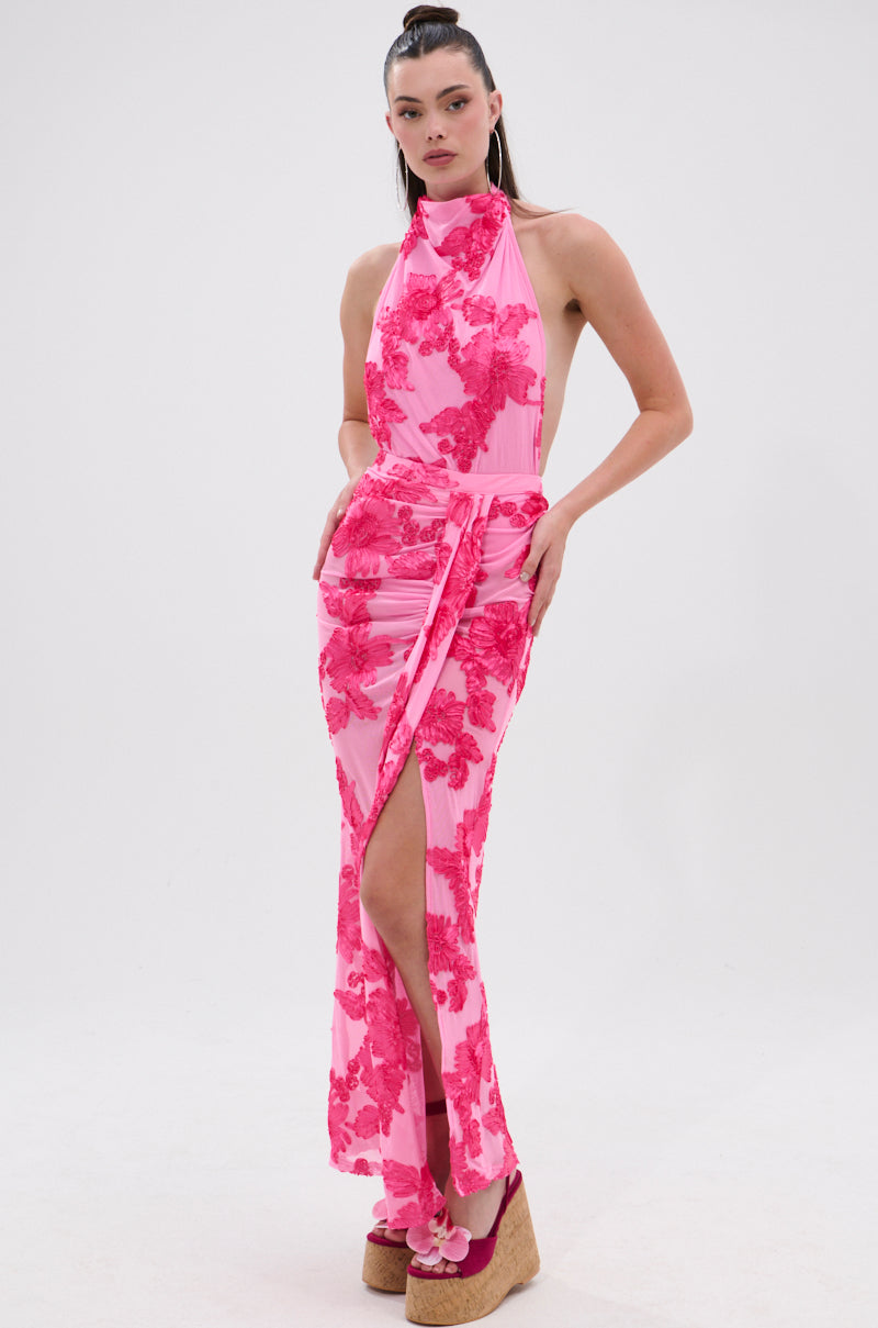 A woman poses in the FLOWER GIRL MESH MAXI SKIRT, featuring satin-textured floral appliqués, a high slit, and a waist tie. She pairs it with wedge sandals and wears her hair in a high ponytail against a plain white background.