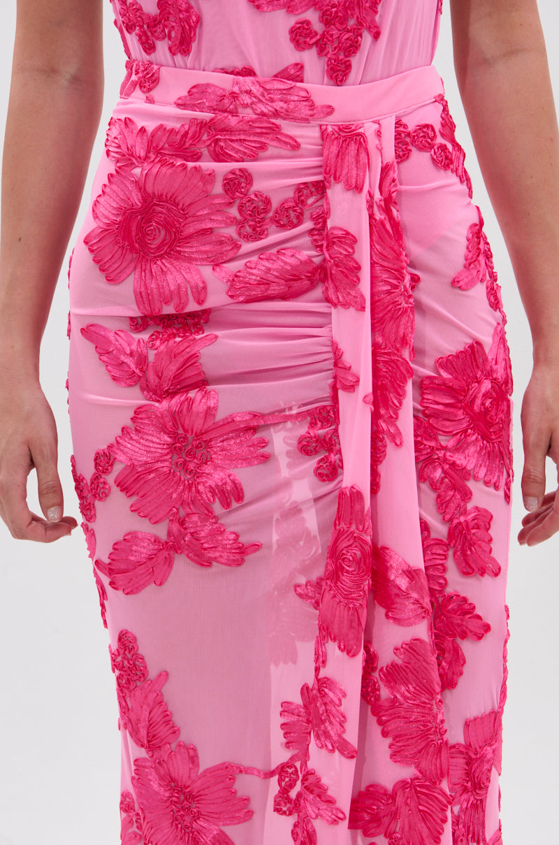 A person wears the FLOWER GIRL MESH MAXI SKIRT, detailed with bright pink satin-textured floral appliqués and a high-rise waist sash, arms relaxed at their sides.