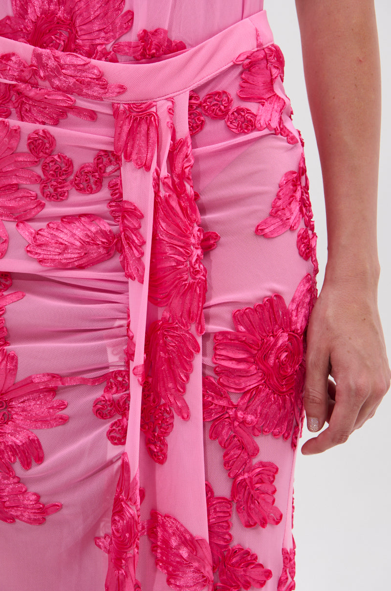 A close-up of a person wearing the FLOWER GIRL MESH MAXI SKIRT, a sheer pink skirt with bright pink satin-textured floral appliqués and a matching sash at the waist. The person's right arm is visible.