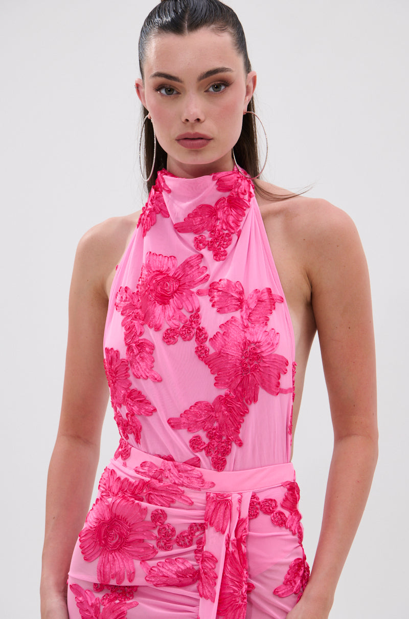 A woman with slicked-back hair wears the FLOWER GIRL MESH BODYSUIT, featuring a sleeveless halter neckline and large, textured dark pink floral appliqués, standing against a plain light background. 1