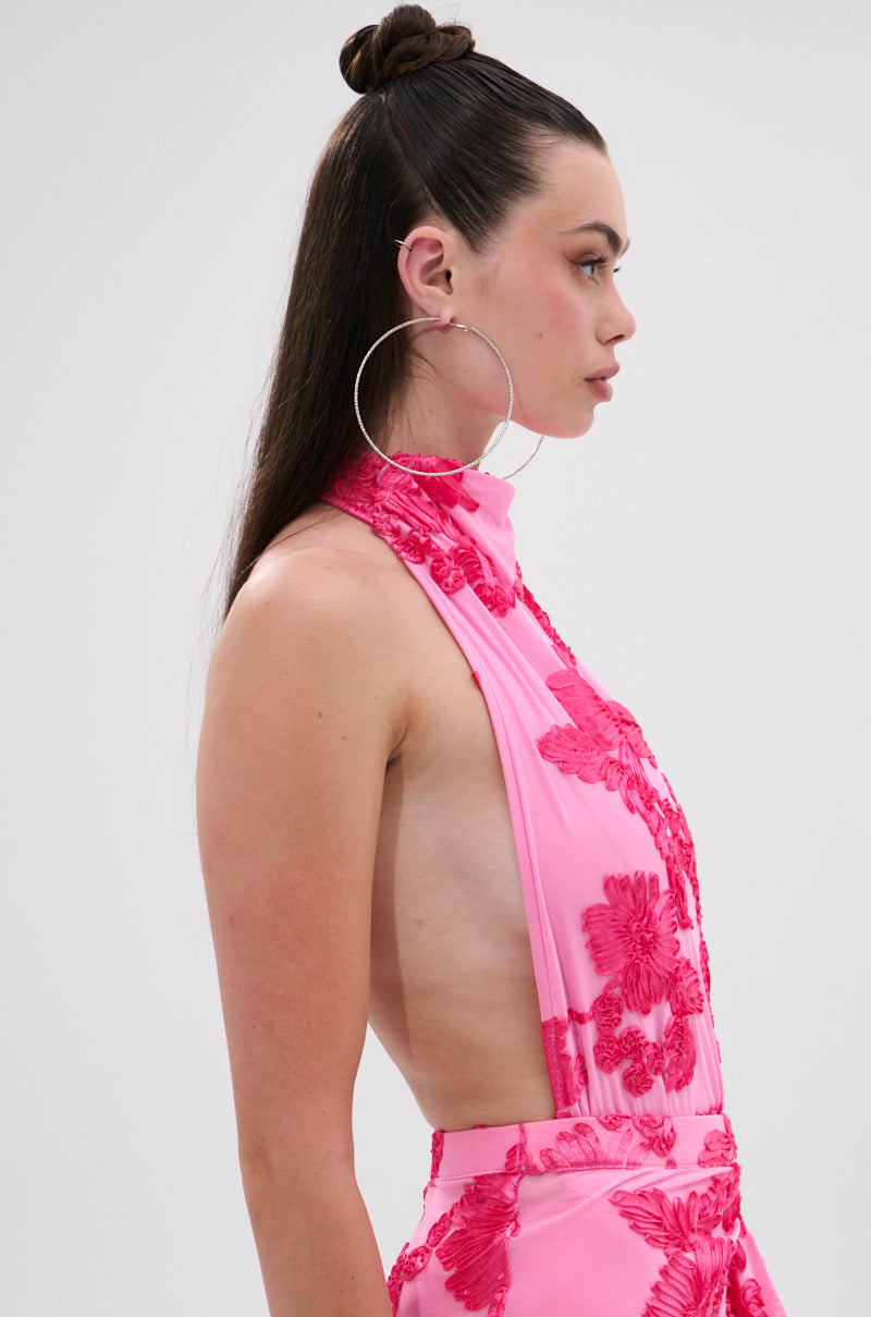 A woman with long dark hair in a high half-bun wears large hoop earrings and the FLOWER GIRL MESH BODYSUIT, a sleeveless, backless pink piece with a halter neckline and red floral embroidery, shown in profile against a plain light background. 2