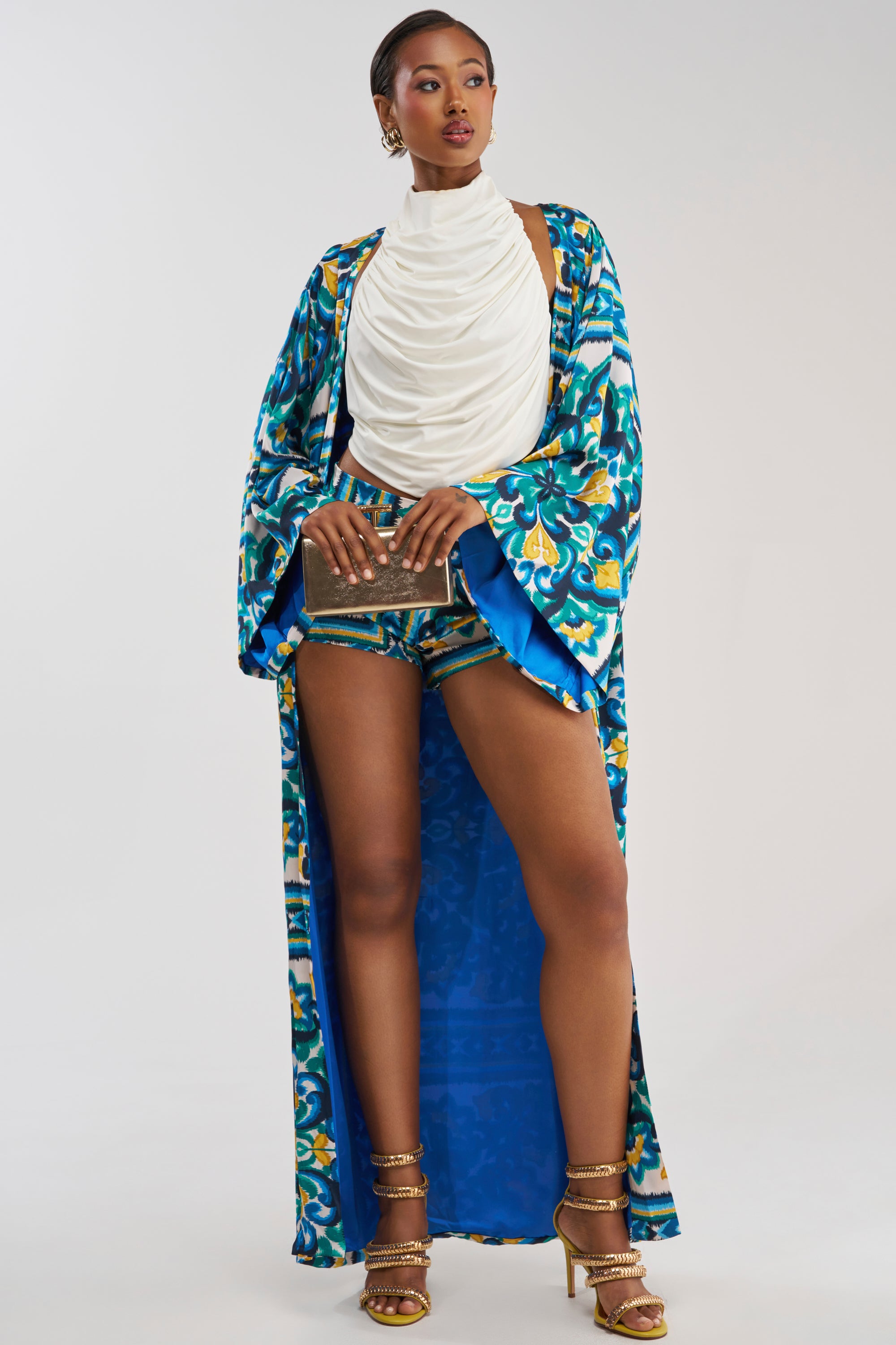 Styled against a plain backdrop, a woman models resort-ready fashion in the FEEL THE HEAT SATIN BLOUSE paired with a blue and green coastal-inspired print robe, matching shorts, gold heels, statement earrings, and a small clutch.