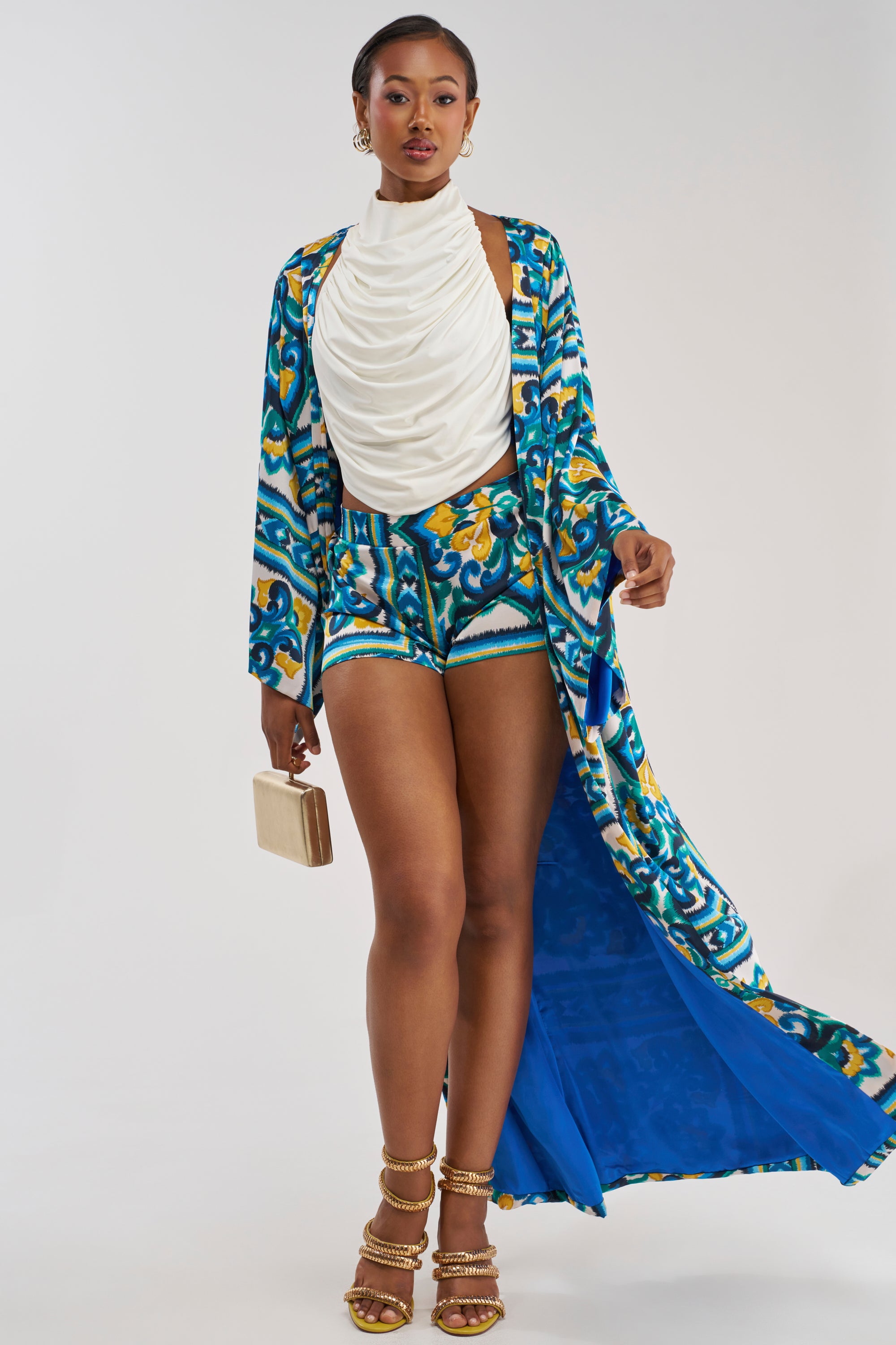 A woman wears the FEEL THE HEAT SATIN BLOUSE, a white draped, coastal-inspired top, paired with a patterned blue and yellow long jacket and matching shorts. She holds a small clutch and accessorizes with gold hoop earrings and heeled sandals against a plain white background.