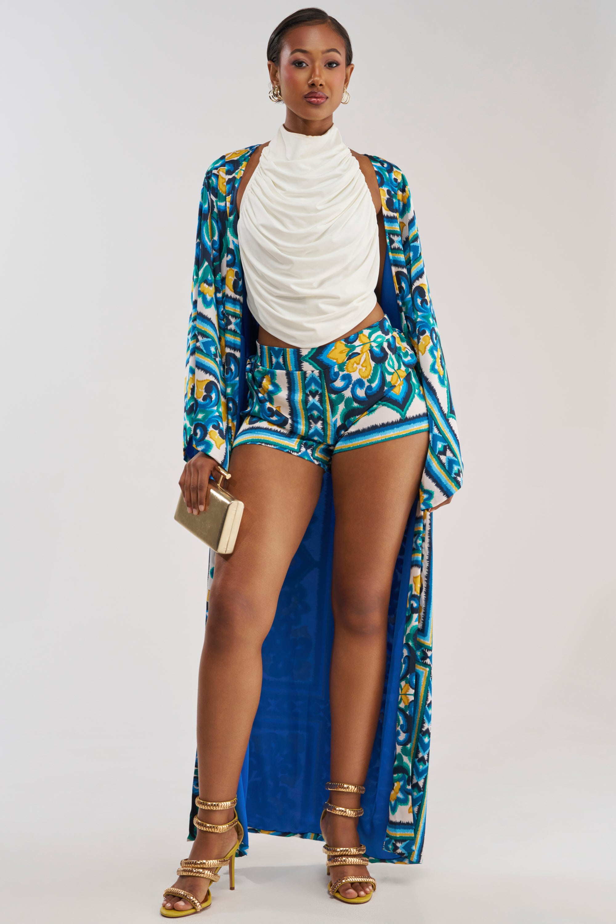 Wearing the FEEL THE HEAT SATIN BLOUSE, a woman poses against a plain background, paired with a coastal-inspired blue, yellow, and white patterned long robe and matching shorts. She completes her look with gold heels, gold earrings, and a small beige clutch.