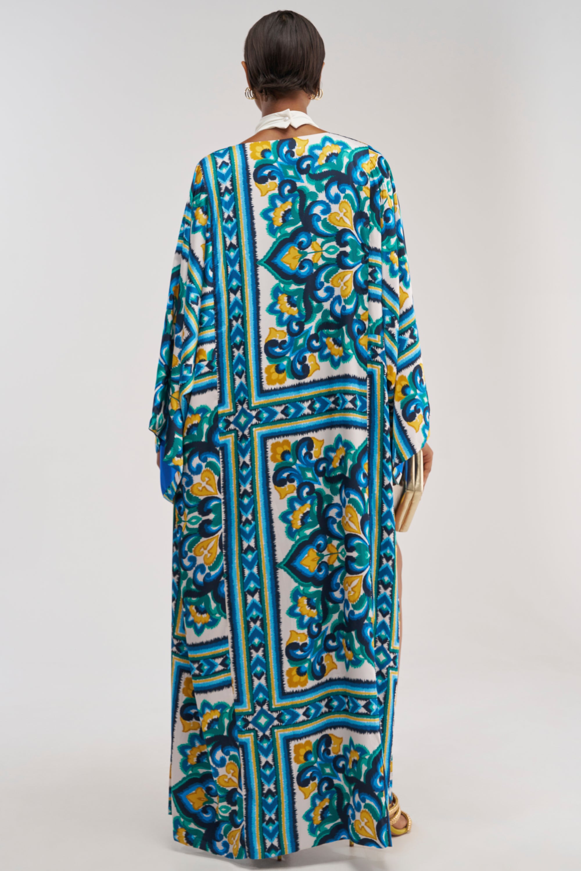 A person stands facing away against a plain light background, wearing the FEEL THE HEAT SATIN BLOUSE, which features long sleeves and vibrant blue, yellow, and green ornate patterns inspired by coastal prints.