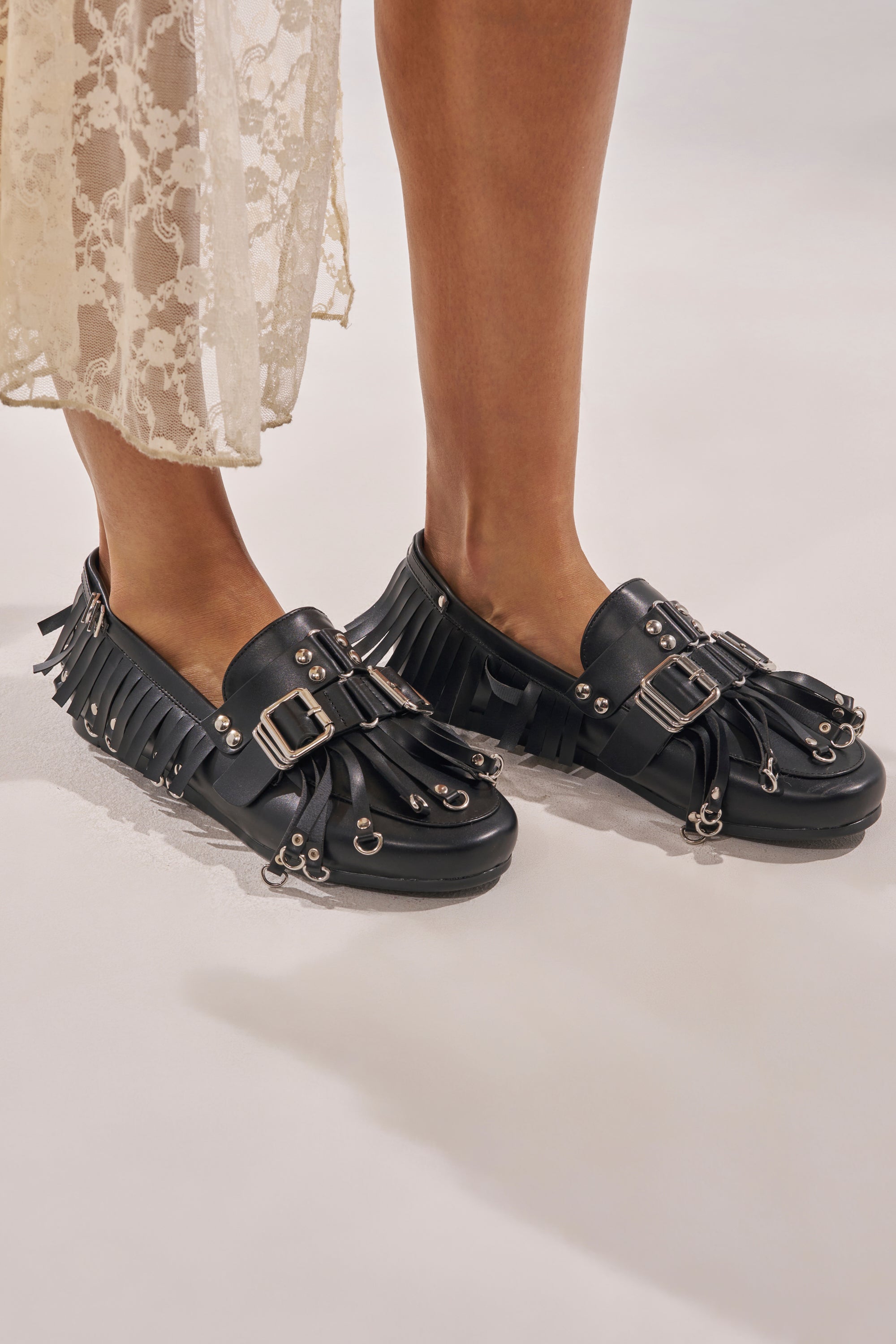 A person stands against a light background in a cream lace skirt and AZALEA WANG SEVYN Black with Silver Hardware Flat Loafers featuring almond toes, silver buckles, studs, fringe, and eyelet accents. 0