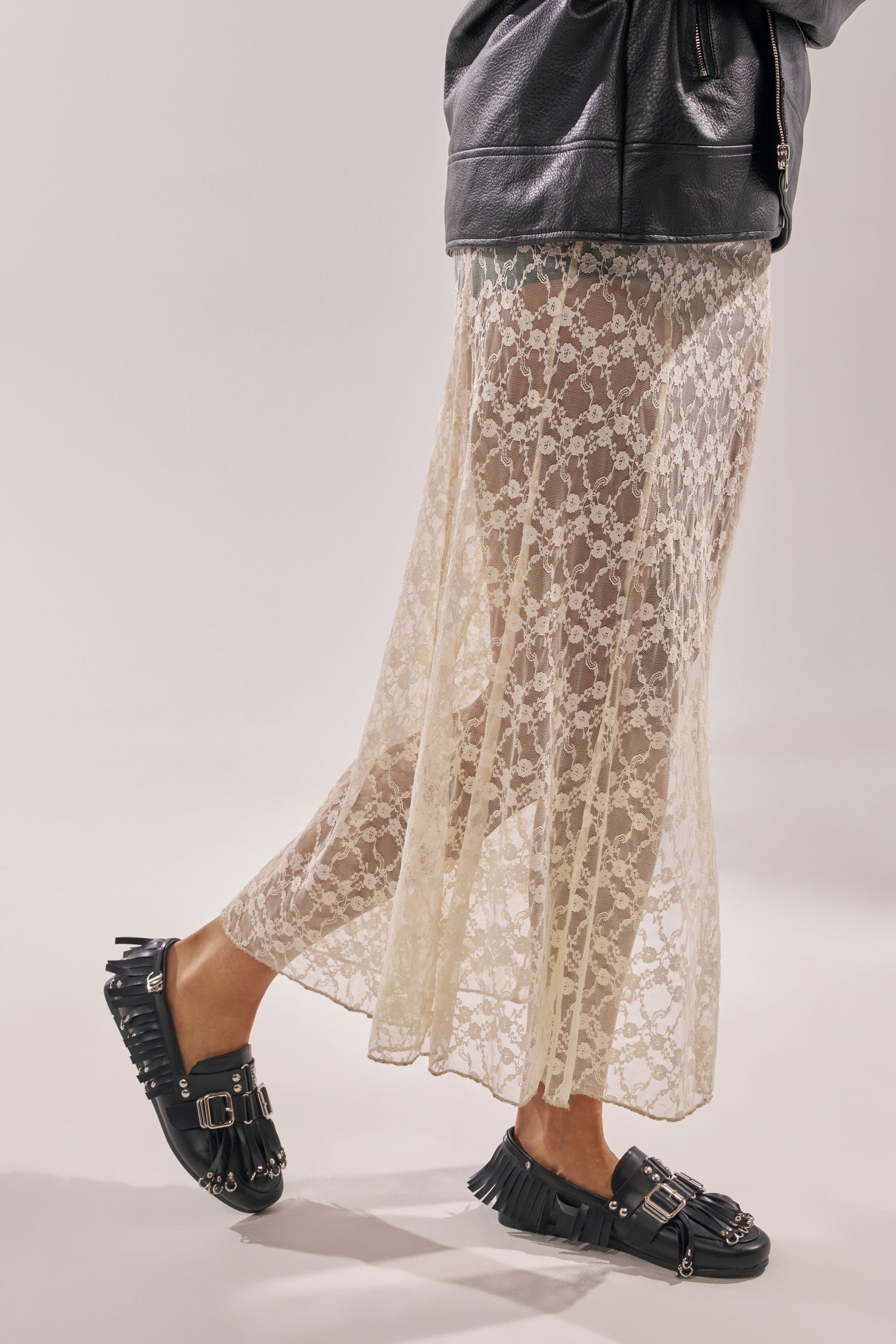 A person in a black leather jacket with silver hardware, a sheer cream lace skirt, and AZALEA WANG SEVYN Black with Silver Hardware Flat Loafers stands against a plain light background. 2