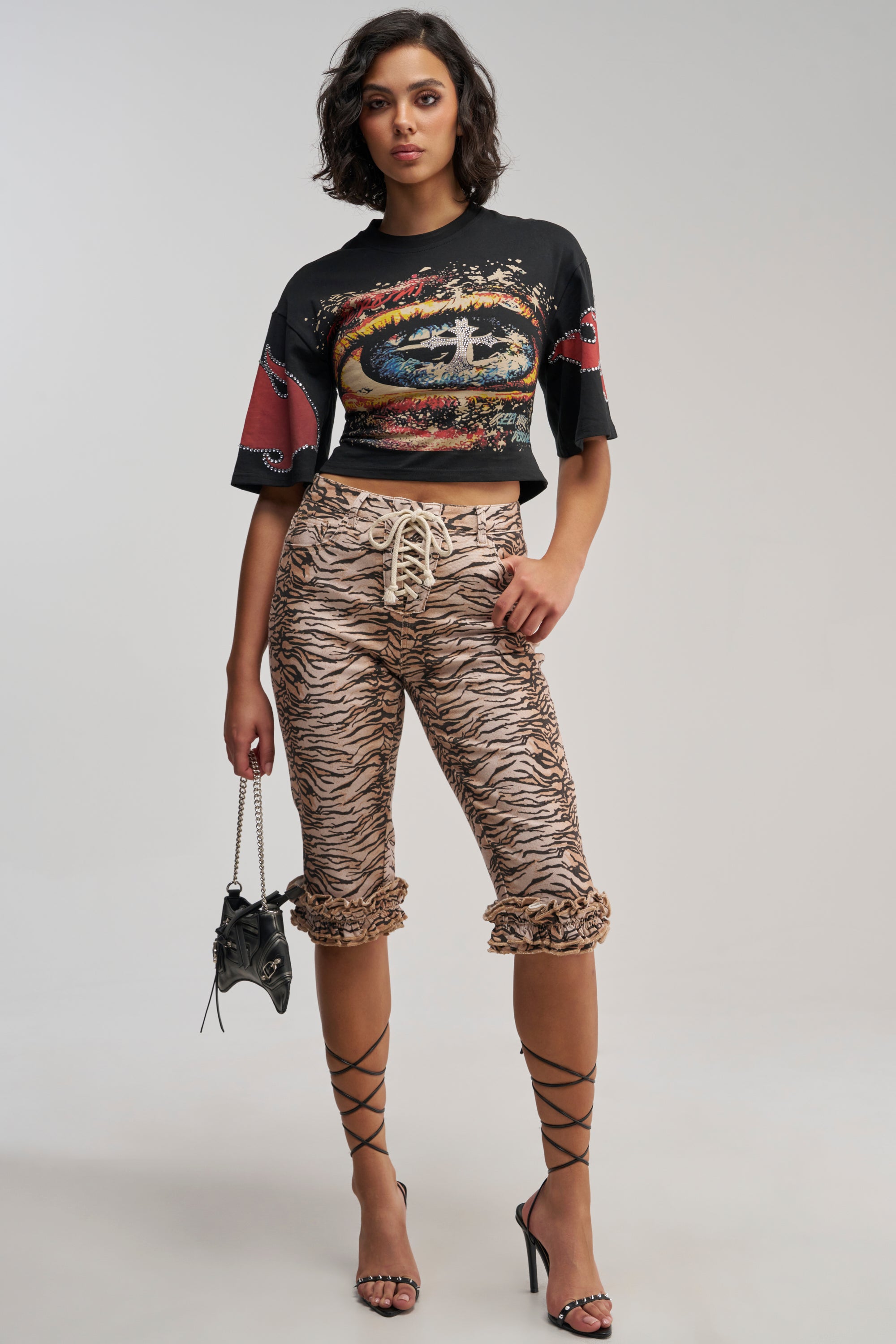 A woman poses in a cropped graphic t-shirt, SEXY TIGER CAPRI pants that are perfect for festival streetwear, strappy black heels, and holds a small black handbag against a plain background.