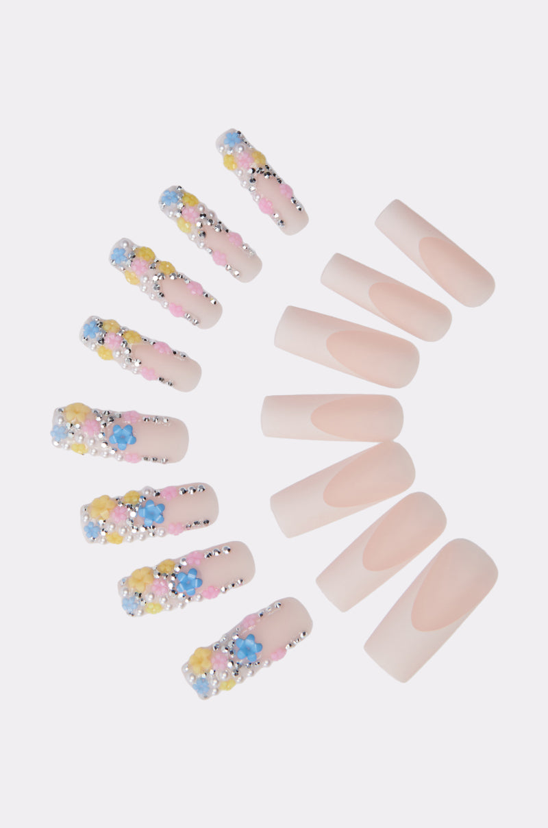 FLOWER POWER FALSE NAILS arranged in a semi-circle on a white background, featuring nude press-ons with curved tips and others decorated with 3D flowers, pastel accents, and rhinestone embellishments.