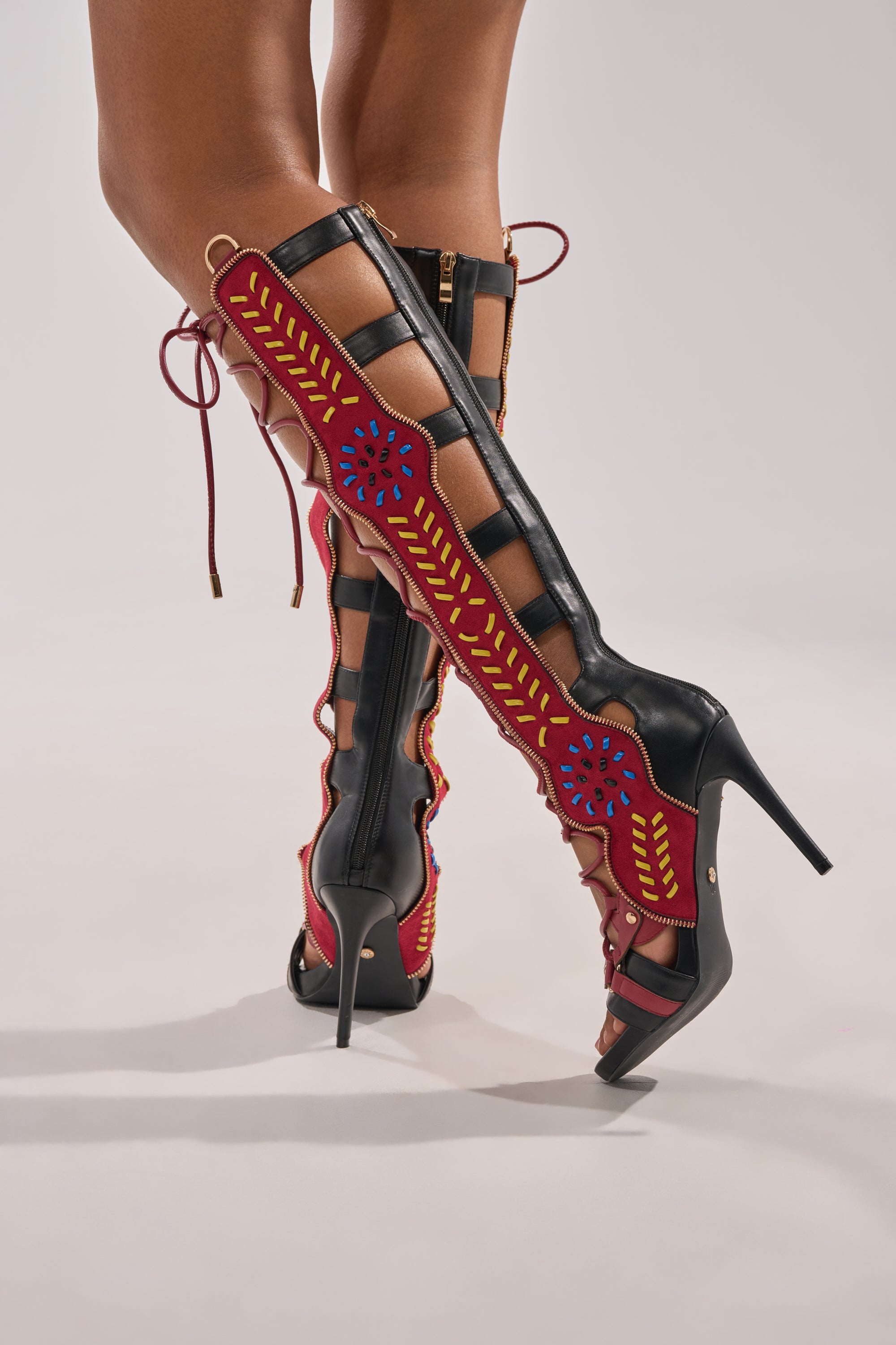 Wearing the AZALEA WANG SHAI BLACK MULTI GLADIATOR SANDAL, featuring black knee-high stilettos with open sides, red trim, yellow leaf designs, and blue floral patterns, a person stands against a plain light background. 0