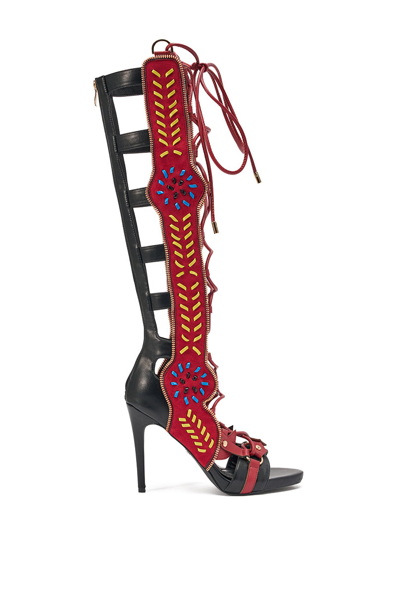 The AZALEA WANG SHAI BLACK MULTI GLADIATOR SANDAL is a knee-high, open-toe stiletto with black straps, a red panel embroidered in yellow and blue, and front lace-up details for a bold statement look.