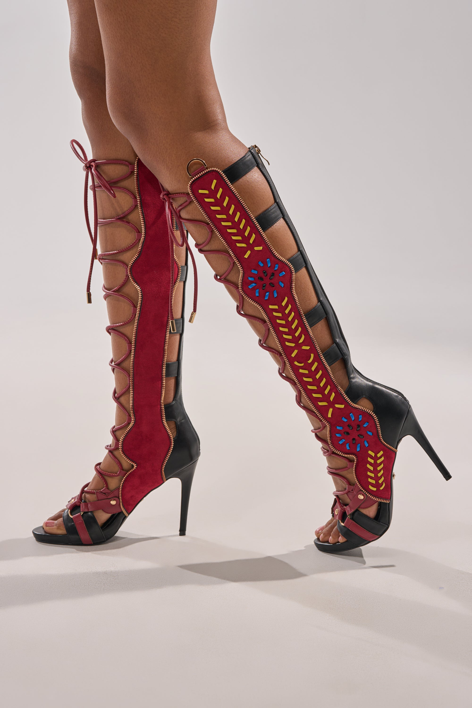 A person wearing AZALEA WANG SHAI BLACK MULTI GLADIATOR SANDALS—knee-high, lace-up stilettos with red, black, and gold accents, side cutouts, decorative stitching, and blue flower designs on the outer sides.