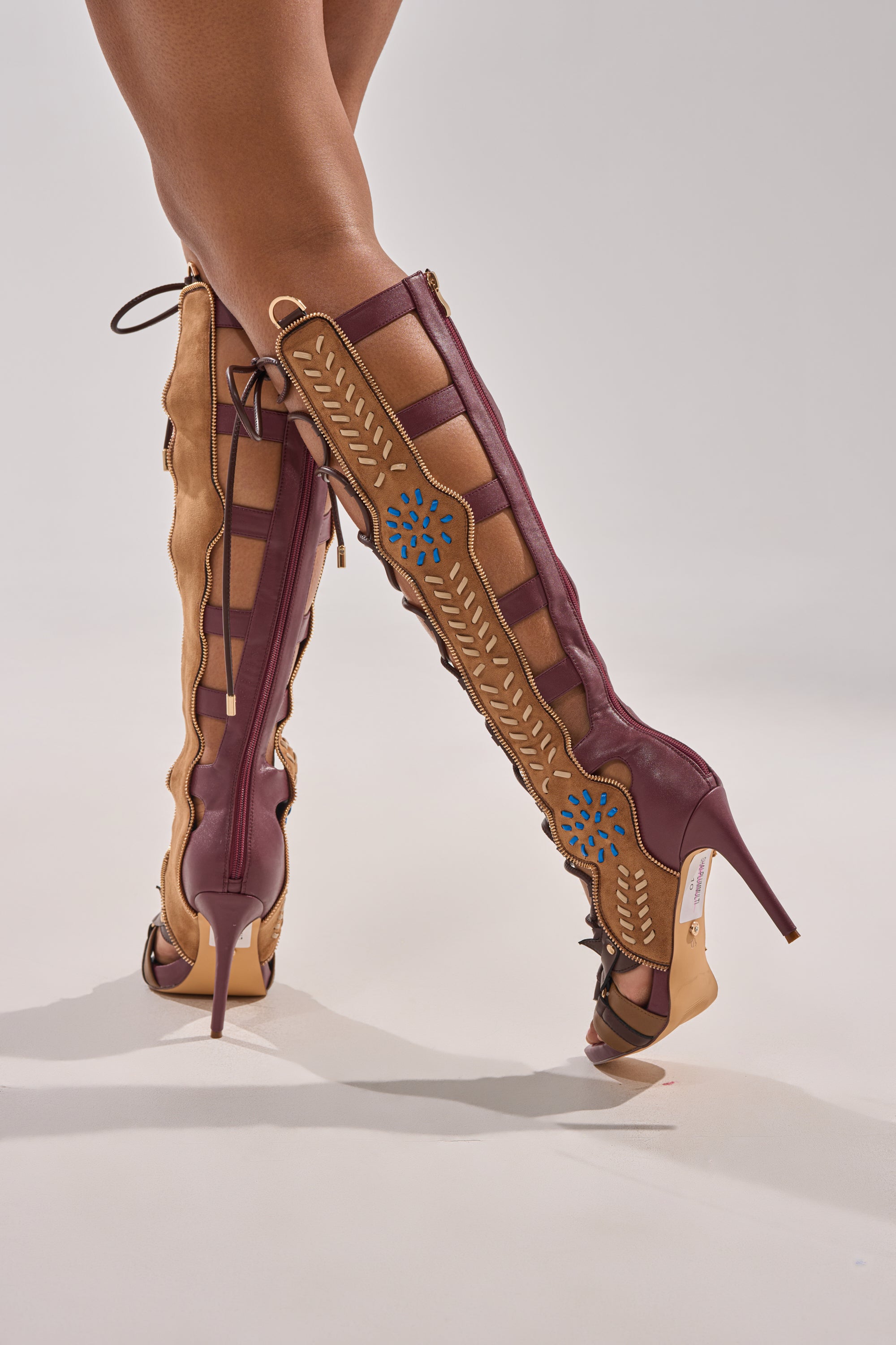 A person stands on tiptoe against a plain background wearing the AZALEA WANG SHAI PLUM MULTI GLADIATOR SANDAL, featuring knee-high tan and burgundy panels, blue floral details, cutouts, and lace-up sides.
