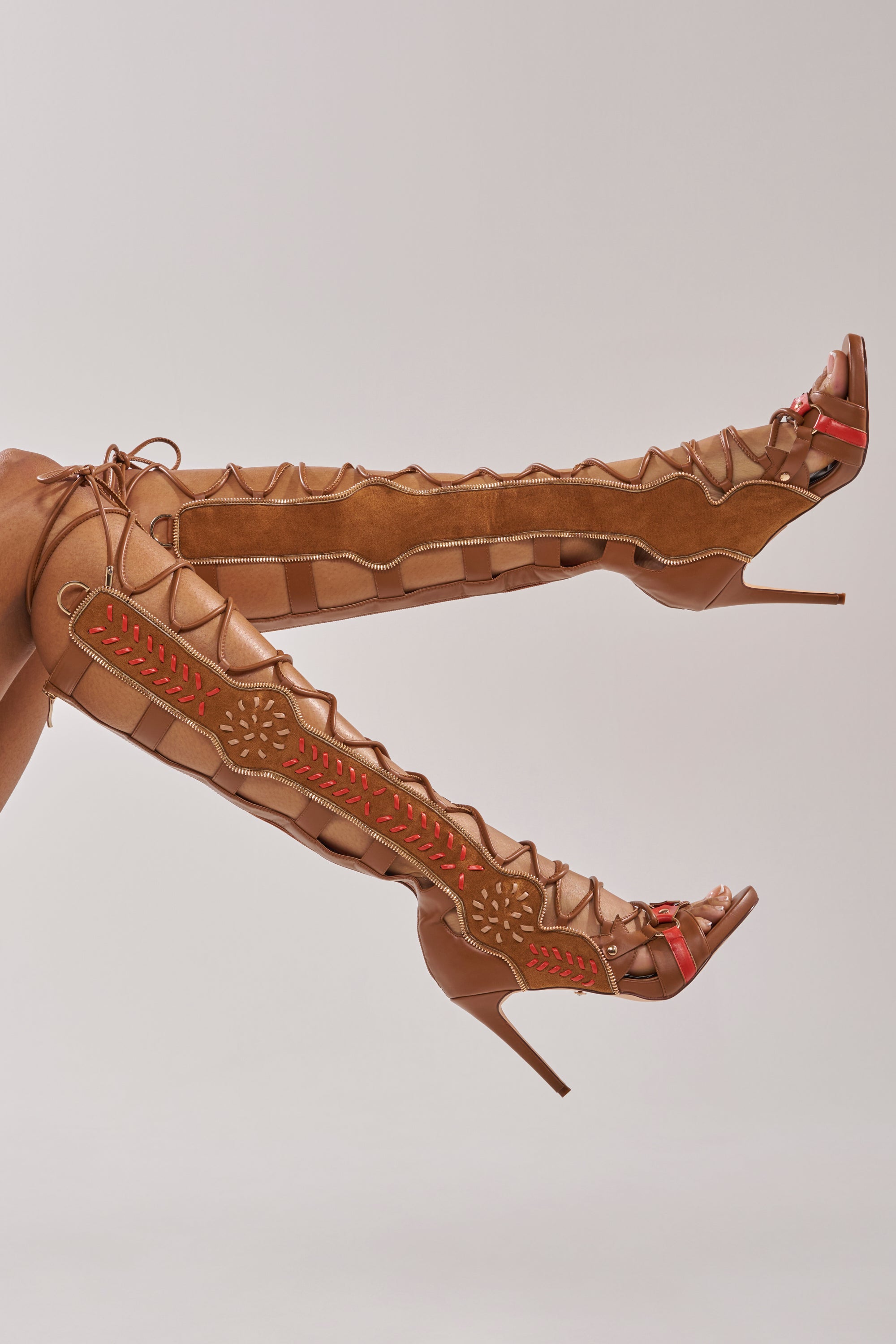 Two legs are raised, showcasing the AZALEA WANG SHAI TAN GLADIATOR STILETTO SANDAL—knee-high heels with intricate red and brown cut-out designs and a tan stiletto silhouette, set against a plain, light background. 1