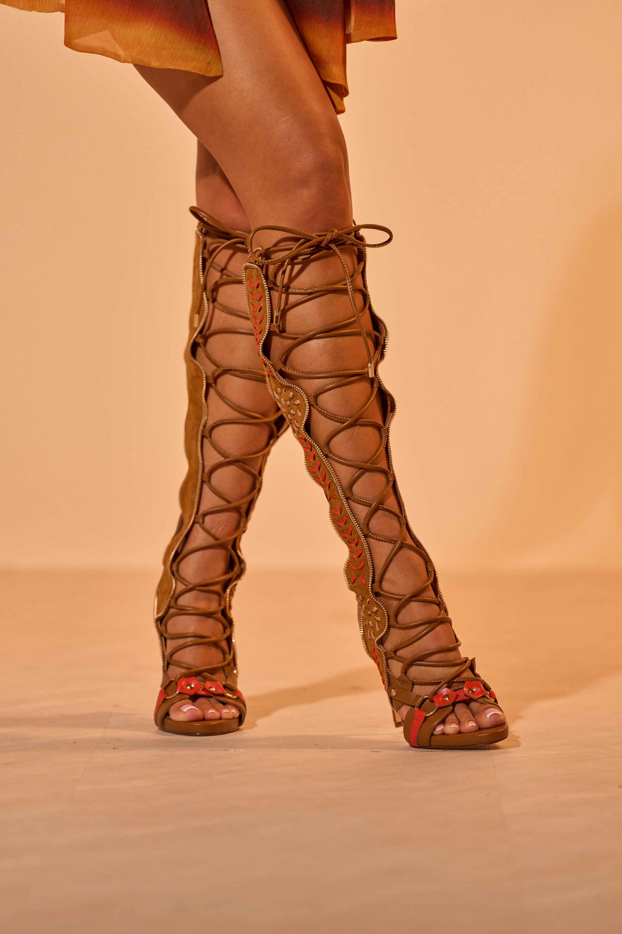 A model showcases the AZALEA WANG SHAI Tan Gladiator Stiletto Sandal, featuring knee-high lace-up styling, open toes, and red accents with a brown and orange dress against a neutral background. 3