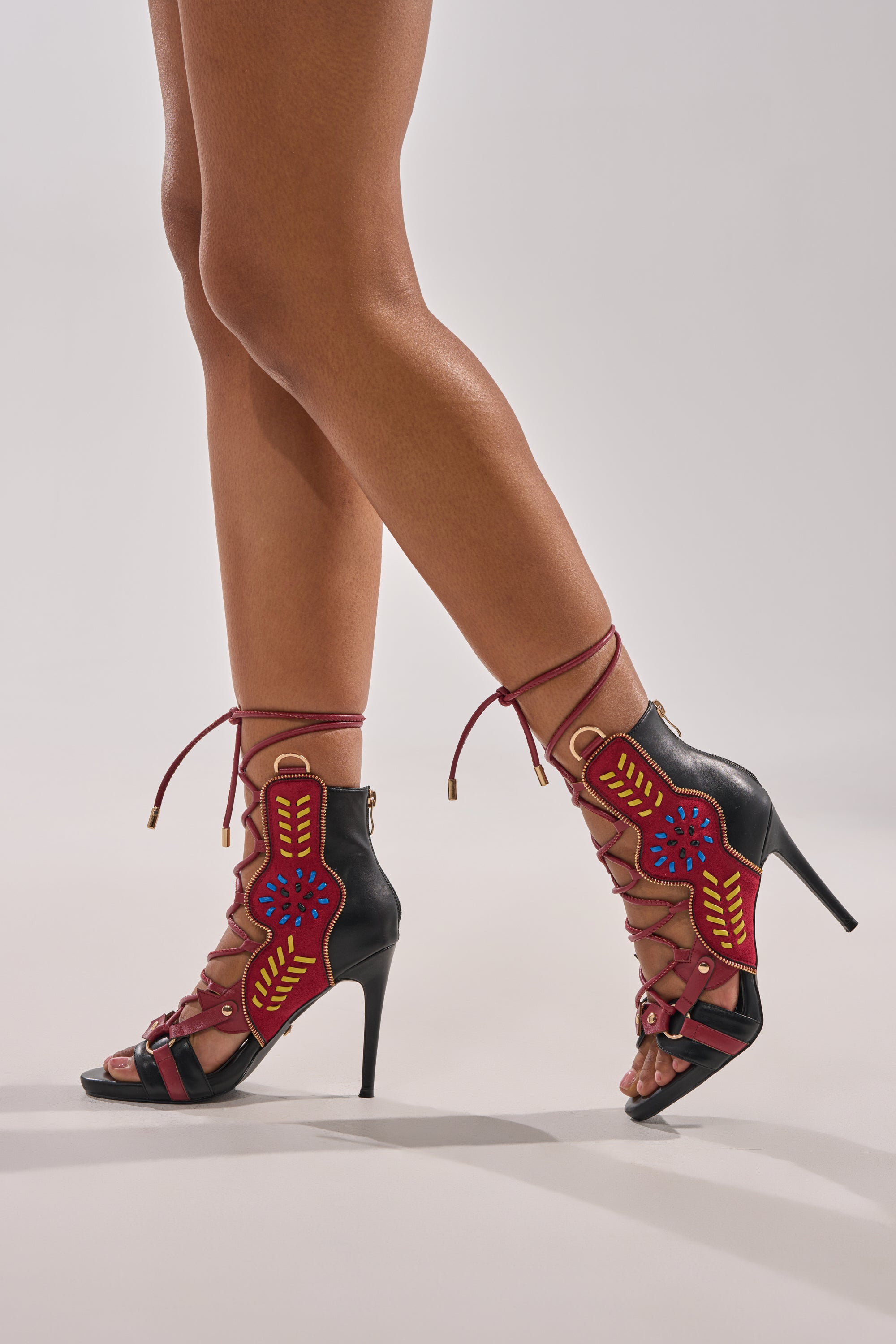 Wearing the AZALEA WANG SHAINIA BLACK MULTI SUEDE STILETTO SANDAL, which features embroidered red, blue, and yellow designs and lace-up straps, a person stands against a plain light background. 1