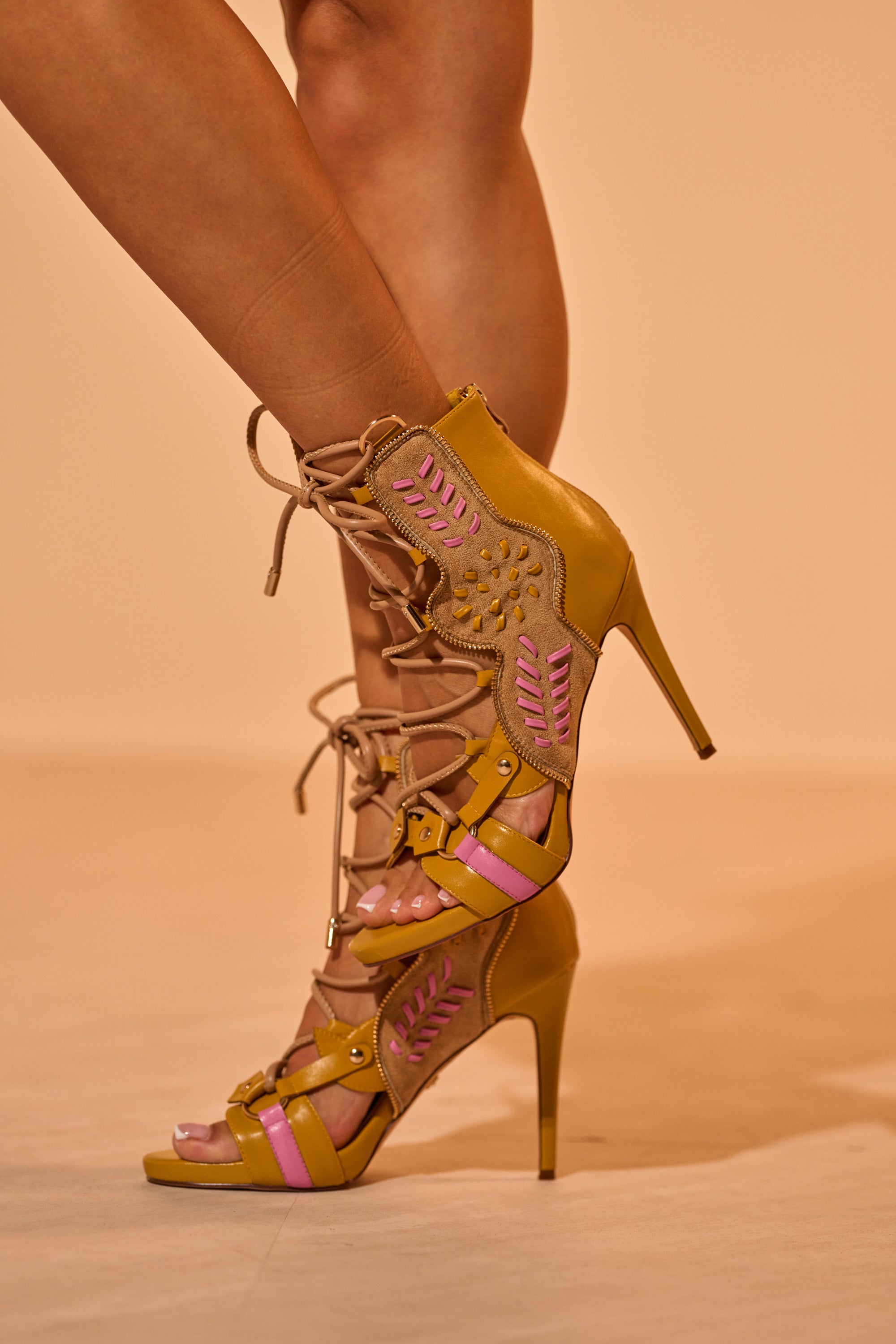 A close-up of legs wearing the AZALEA WANG SHAINIA MUSTARD MULTI STILETTO SANDAL, featuring mustard yellow and pink accents, lace-up ankle details, stylish suede paneling, and intricate cutout designs. 5