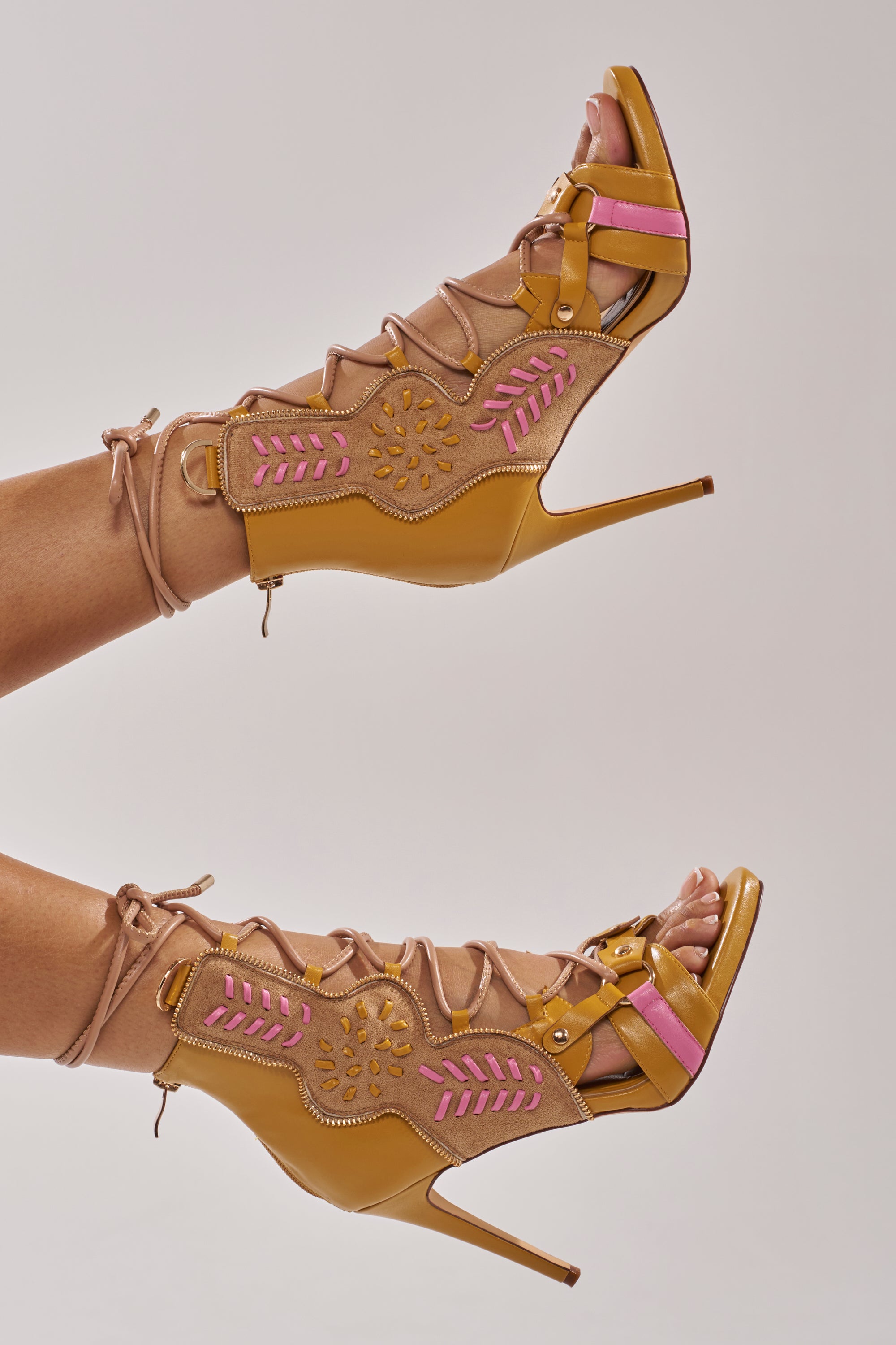 The AZALEA WANG SHAINIA Mustard Multi Stiletto Sandal, featuring tan lace-up gladiator style, cutouts, pink and yellow accents, and floral patterns, is worn on feet against a plain light background. 7