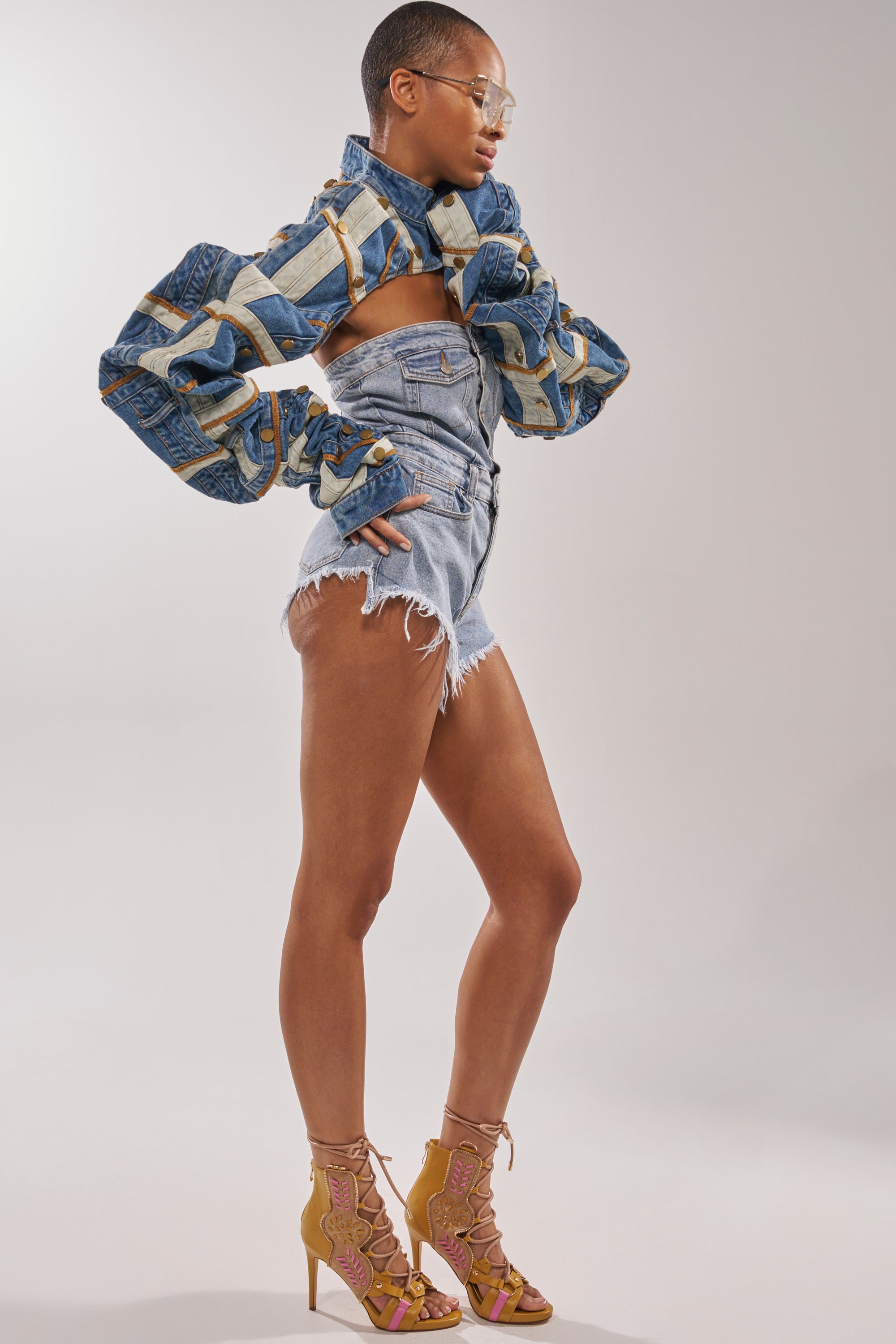 A person wearing a patchwork denim jacket, high-waisted distressed denim shorts, clear glasses, and the AZALEA WANG SHAINIA MUSTARD MULTI STILETTO SANDAL poses confidently against a plain background. 10