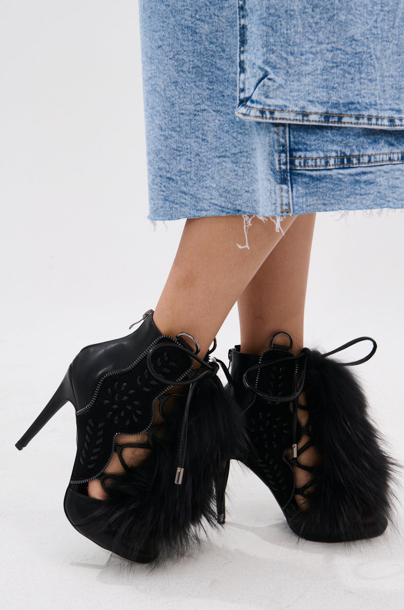A person wears a frayed light blue denim skirt and the AZALEA WANG SHAINY BLACK FUR SANDAL, which features bold cutout patterns and faux fur details for a standout look.
