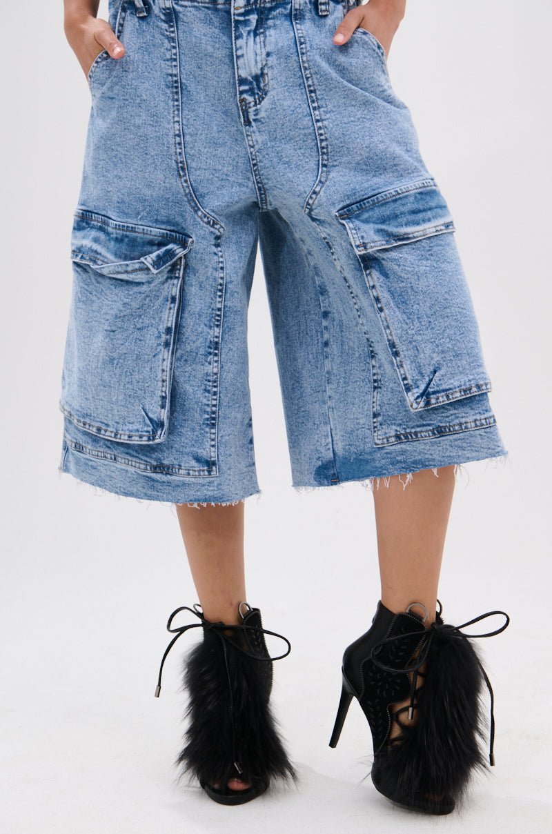 A person wears knee-length, light-wash denim shorts with large front pockets and frayed hems, styled with AZALEA WANG SHAINY BLACK FUR SANDALS featuring stiletto heels, fur accents, and lace-up details.