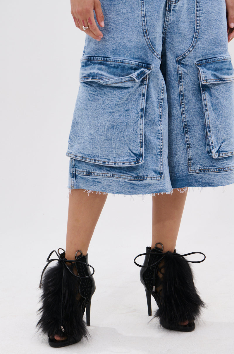 A person wearing light blue, knee-length denim shorts with large front pockets and frayed hems, paired with the AZALEA WANG SHAINY BLACK FUR SANDAL featuring lace-up fronts and faux fur accents.