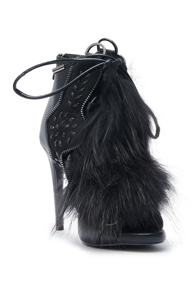 The AZALEA WANG SHAINY BLACK FUR SANDAL is a stylish faux leather high-heeled sandal with zipper details, decorative cutouts, and a bold black fur accent on the side.