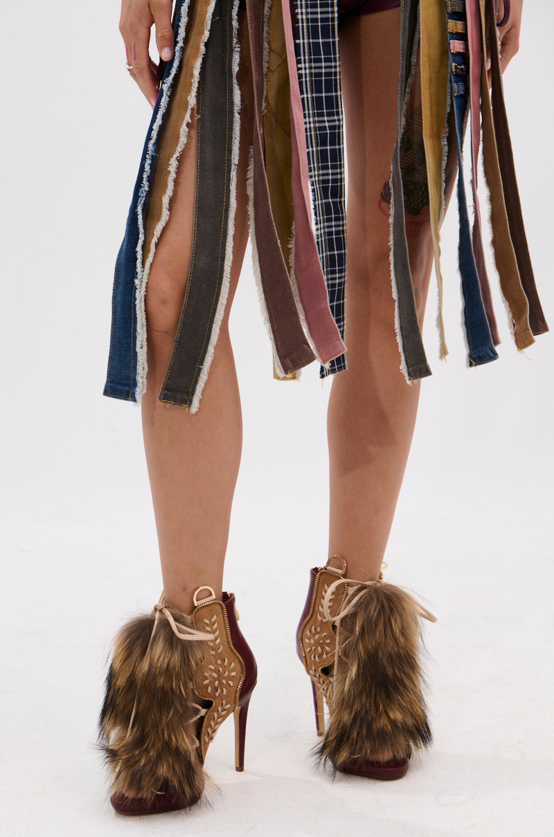 A person wears the AZALEA WANG SHAINY MULTI MATERIAL SANDAL, paired with an outfit featuring multicolored, frayed fabric strips hanging to the knees.