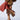 A person is wearing a red leather skirt, and a patterned coat with fur trim. They have on high-heeled shoes featuring stiletto heels and lace-up details, specifically the AZALEA WANG SHAINY MULTI MATERIAL SANDAL. In their hand, they hold one shoe. The background is plain gray.
